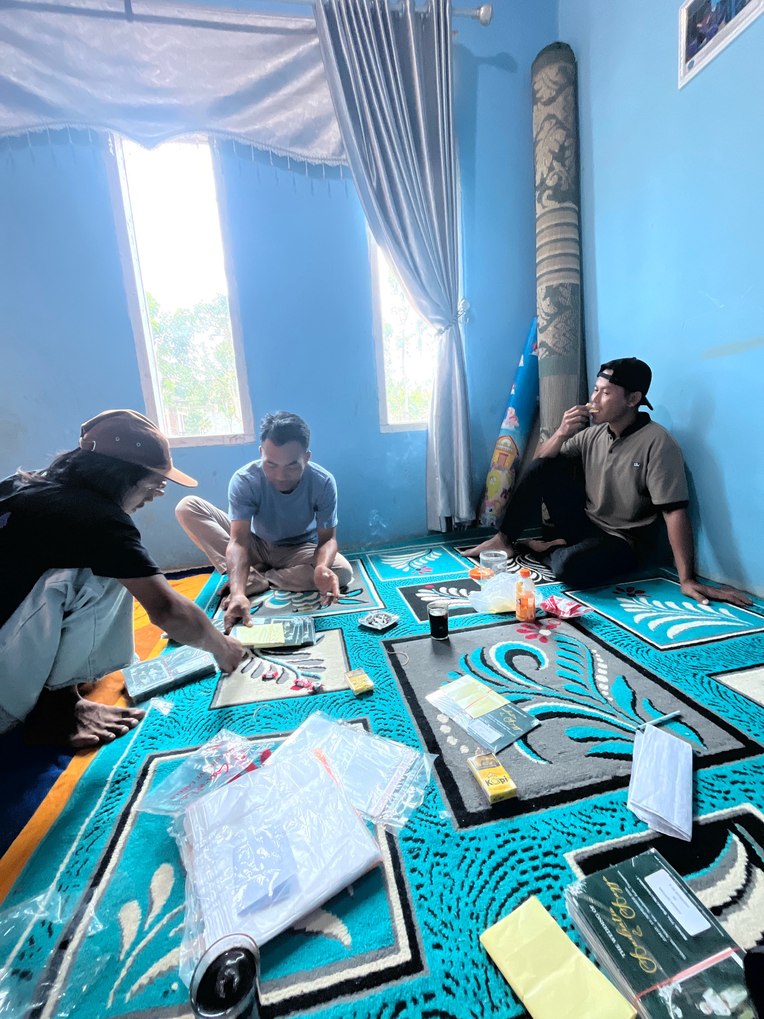 The image depicts an indoor scene in Kebumen, Indonesia, where three men are gathered on the floor, which is covered with colorful rugs. Two men are seated on the floor, facing each other, engaged in an activity that involves placing items on a rug. One man is wearing a black t-shirt and a brown baseball cap, and he is leaning forward. The other man is wearing a light blue t-shirt and light-colored pants, and he is holding a cigarette. A third man is seated on the floor to the right, wearing a brown polo shirt and a black baseball cap, and he is eating or drinking. The room has light blue walls and two windows with light-colored curtains. Various objects are scattered on the rugs, including plastic packaging, cigarette packs, a bottle of drink, and what appear to be cards or documents. The overall impression is a casual gathering in a domestic setting.