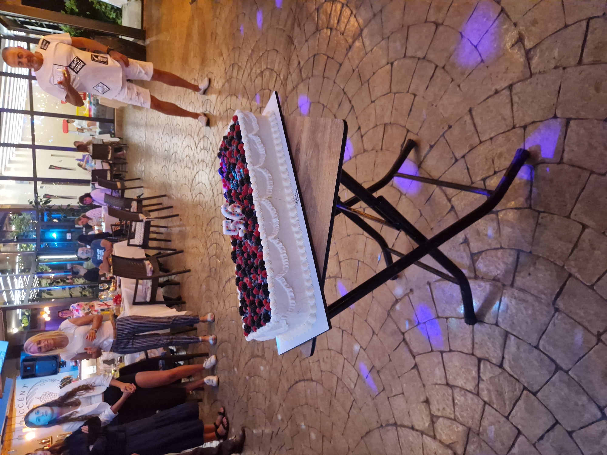 An outdoor paved patio in Tankovo, Bulgaria, is the setting for a celebration, likely a birthday, given the cake. The cake, a rectangular white frosted confection topped with a dense arrangement of raspberries and blackberries, features two large candle numbers, "5" and "0", indicating a 50th birthday. The cake rests on a dark, collapsible table with a wooden top, positioned on the patterned stone ground. In the background, several individuals are present. On the left, a woman in a dark dress and sandals stands next to another woman in a white top and black skirt. To the right, a man in a white t-shirt with graphic patches and white distressed shorts holds a glass and appears to be the focus of attention. Other guests are visible seated at tables in the background, some of whom are dining or socializing. The overall ambiance suggests a festive outdoor gathering.