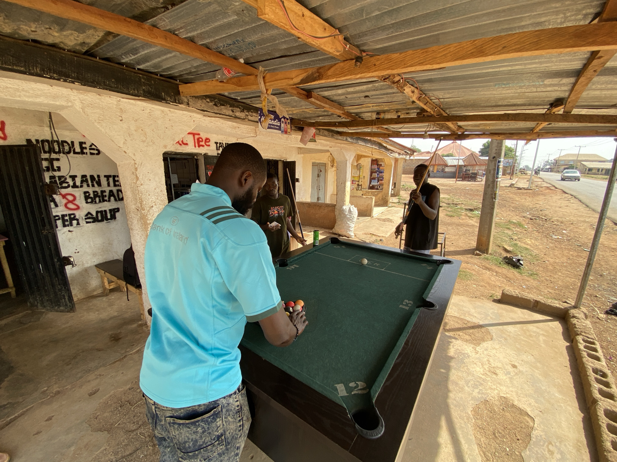 This image captures a vibrant outdoor scene in Vom, Nigeria, under a brightly lit, likely mid-day sun, indicated by strong shadows and clear skies. The setting appears to be a rustic, open-air establishment, possibly a shop or game center, covered by a corrugated metal roof supported by rough-hewn wooden beams. The ground is a mix of concrete and dirt.
Three men are present, gathered around a green pool table. In the foreground, a man with a beard, wearing a light blue polo shirt with "Bank of Ireland" text and logo, and faded jeans, is bent over the pool table. He is in the process of arranging the colored pool balls into a rack, suggesting the start or restart of a game. A white cue ball rests further down the table, near partially visible text that includes "EN". The pool table itself has numbers "13" and "12" printed on its surface near two pockets.
Behind him, to the left, another man in a dark t-shirt with "JUST DO IT" partially visible text, stands observing. To the right, a third man in a dark sleeveless shirt holds a pool cue, ready to play or watch.
The background reveals a whitewashed wall with hand-painted black and red text advertising items for sale: "NOODLES", "EGG", "ARABIAN TEA", "BREAD", and "PEPPER SOUP". Another snippet reads "Tee" (likely part of "Tea"). Beyond the structure, a dusty road is visible with a car in the distance, and simple buildings characteristic of a semi-urban or rural Nigerian landscape. Visible objects include empty plastic bottles, a white sack, and a wooden bench. The overall atmosphere suggests a casual, social gathering.