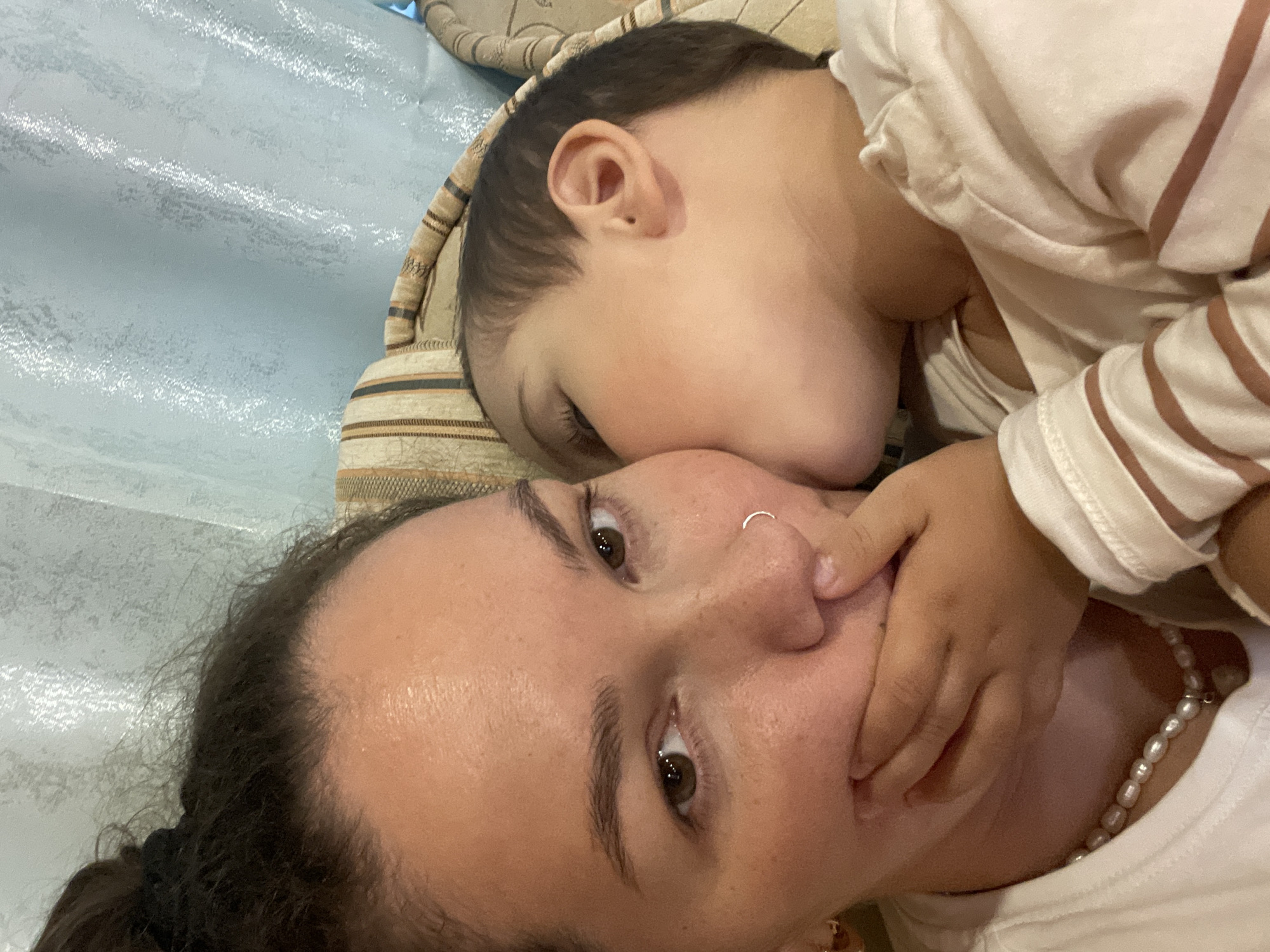 This close-up, overhead shot captures an intimate moment between a mother and her child. The mother's face, tilted upwards, occupies the foreground. She has dark hair pulled back, brown eyes, and a subtle nose ring. Her expression appears calm and affectionate, as she looks directly at the camera. A small child is nestled against her cheek, with their face turned away from the camera. The child's hand is gently touching the mother's face, and their head is resting on a striped cushion. Both mother and child are wearing light-colored clothing. The mother is wearing a pearl necklace. The background is a soft, textured surface, possibly a bed or couch, with a hint of light blue and beige tones. The overall mood is one of tenderness and connection. The lighting suggests it could be daytime. There is no visible text in the image.