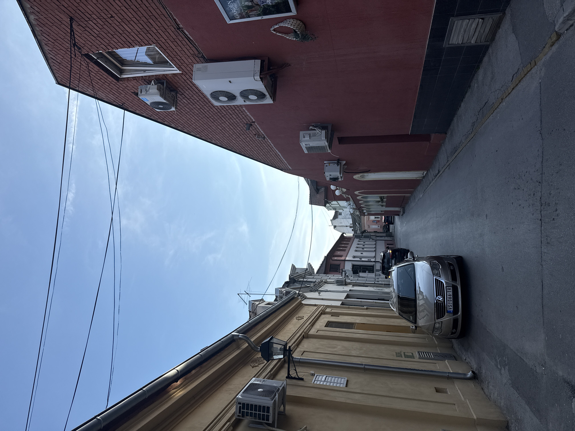 A low-angle shot of a narrow street in an urban area. The street is lined with buildings of varying heights and architectural styles, painted in different colors. On the left, a building with a light-colored facade has an air conditioning unit mounted on its wall. The building on the right has a reddish-brown facade with multiple air conditioning units installed near the top. Electrical wires stretch across the sky. A car is parked on the right side of the street, facing the camera. Other cars and buildings are visible further down the street. The sky is blue with some clouds.