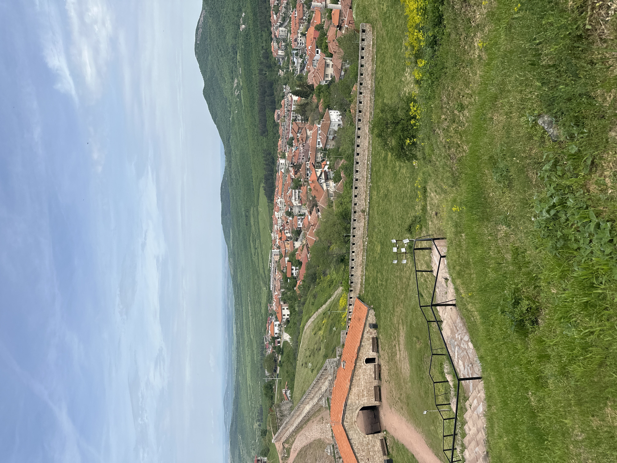 The image depicts a high-angle view of Belogradchik, Bulgaria. The foreground showcases a stone fortress wall with orange-tiled roofs, leading down a grassy slope traversed by a stone-paved path lined with metal railings. Beyond the fortress, the town of Belogradchik stretches into the distance, its buildings clustered together with orange-tiled roofs amidst green trees. A densely forested mountain rises steeply behind the town, creating a dramatic backdrop. The sky above is partly cloudy.