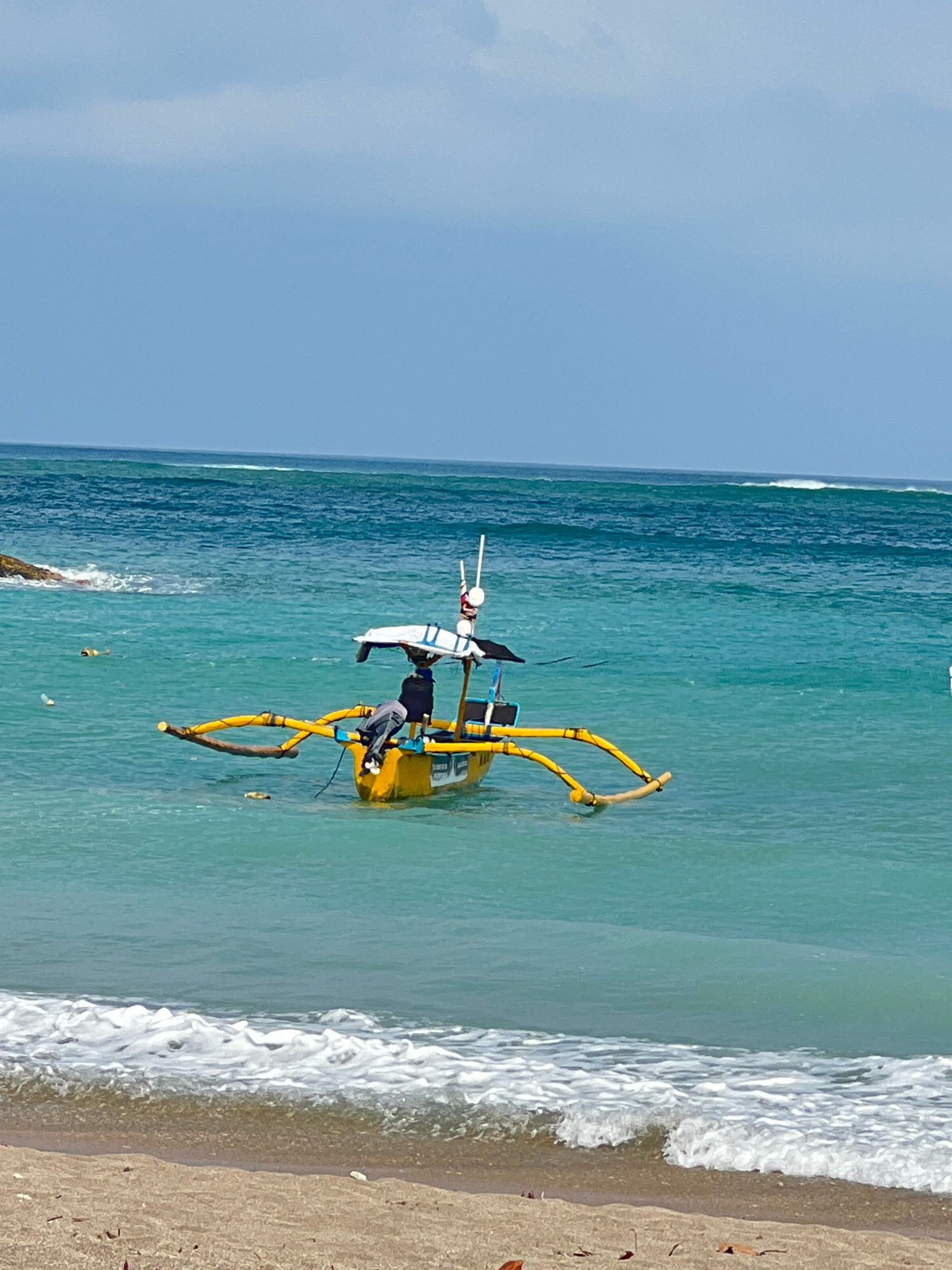 A traditional Balinese outrigger boat, painted bright yellow, floats on the turquoise waters off the coast of Kuta, Indonesia. The boat has a simple canopy providing shade for a person sitting in it. The outriggers, made of bamboo, extend outwards from the main hull. In the background, gentle waves break on the shore, and the sky is a light blue with some clouds. The beach in the foreground is sandy with some scattered debris. The scene suggests a tranquil day at sea.