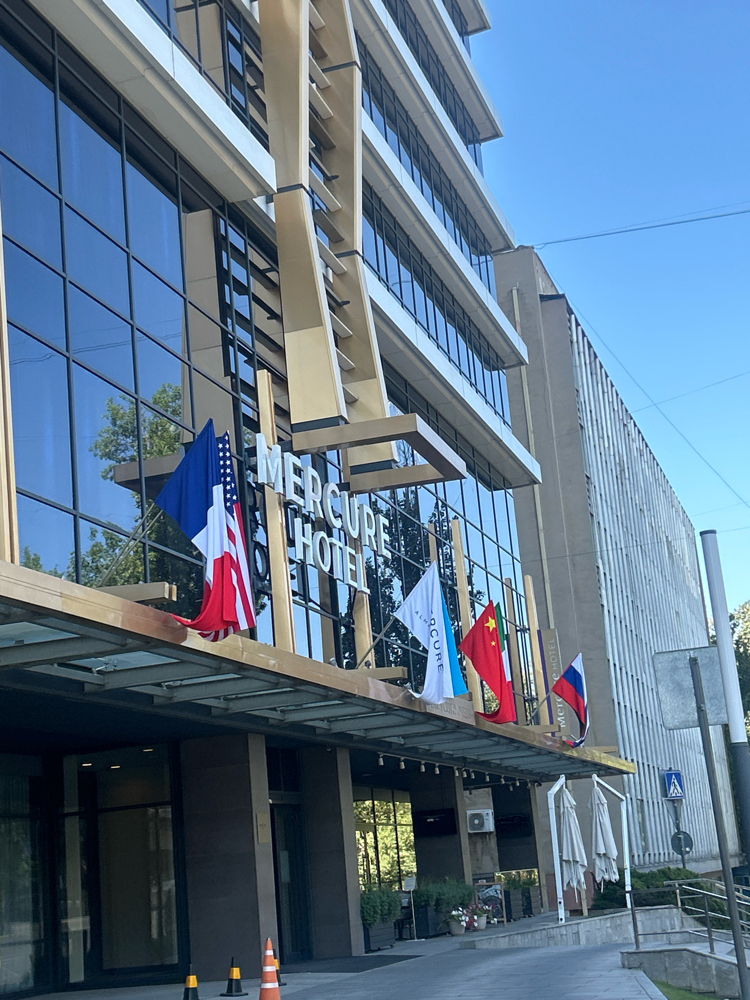 The image shows the exterior of the Mercure Hotel in Almaty, Kazakhstan, on a clear, sunny day. The hotel is a modern building with a glass facade and beige architectural elements. Flags of several countries, including the United States, France, China, and Russia, are displayed in front of the hotel entrance. The entrance area features a covered walkway, potted plants, and outdoor seating. In the foreground, traffic cones are placed on the pavement. The building is surrounded by trees and a street with a pedestrian crossing sign. The overall scene depicts a well-maintained hotel entrance in an urban setting.