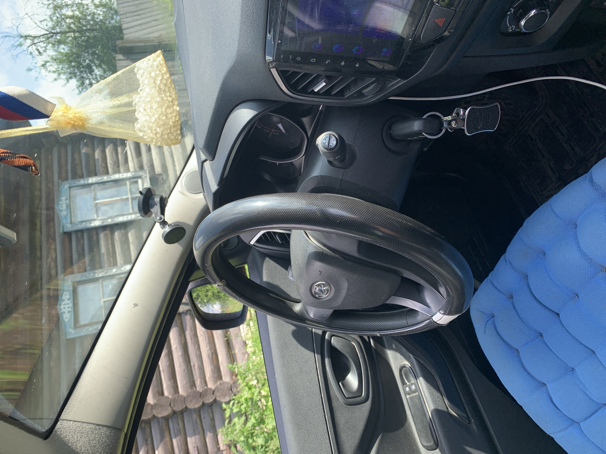 This media file shows the interior of a car parked outside a wooden house in what appears to be a rural setting in Barsai, Russia. The sunlight suggests it is daytime, possibly morning or afternoon.
The primary subjects are the car's interior elements: a steering wheel, dashboard with a touch screen display, car keys in the ignition, and a portion of a blue upholstered seat. Hanging from the rearview mirror or similar attachment point on the windshield is a decorative item consisting of a ribbon with red, white, and blue stripes and a small, sheer golden pouch filled with what looks like small, pearl-like beads. A dark fabric ribbon with an orange and black pattern is also visible, possibly a St. George's ribbon, often associated with patriotic or remembrance events.
The scene is viewed from the perspective of someone sitting inside the car, looking towards the dashboard and passenger side. Through the windshield, a rustic wooden house with two windows featuring decorative white trim is visible. The exterior appears to be a sunny day with some greenery and trees in the background, suggesting a natural, possibly summer environment.
There is no visible text in the image, except for what might be on the car's display screen, which is too blurry to decipher. The overall impression is a snapshot of a car interior on a bright day, with elements that hint at a cultural context within Russia. No people are visible, and no specific activity is taking place other than the car being parked.