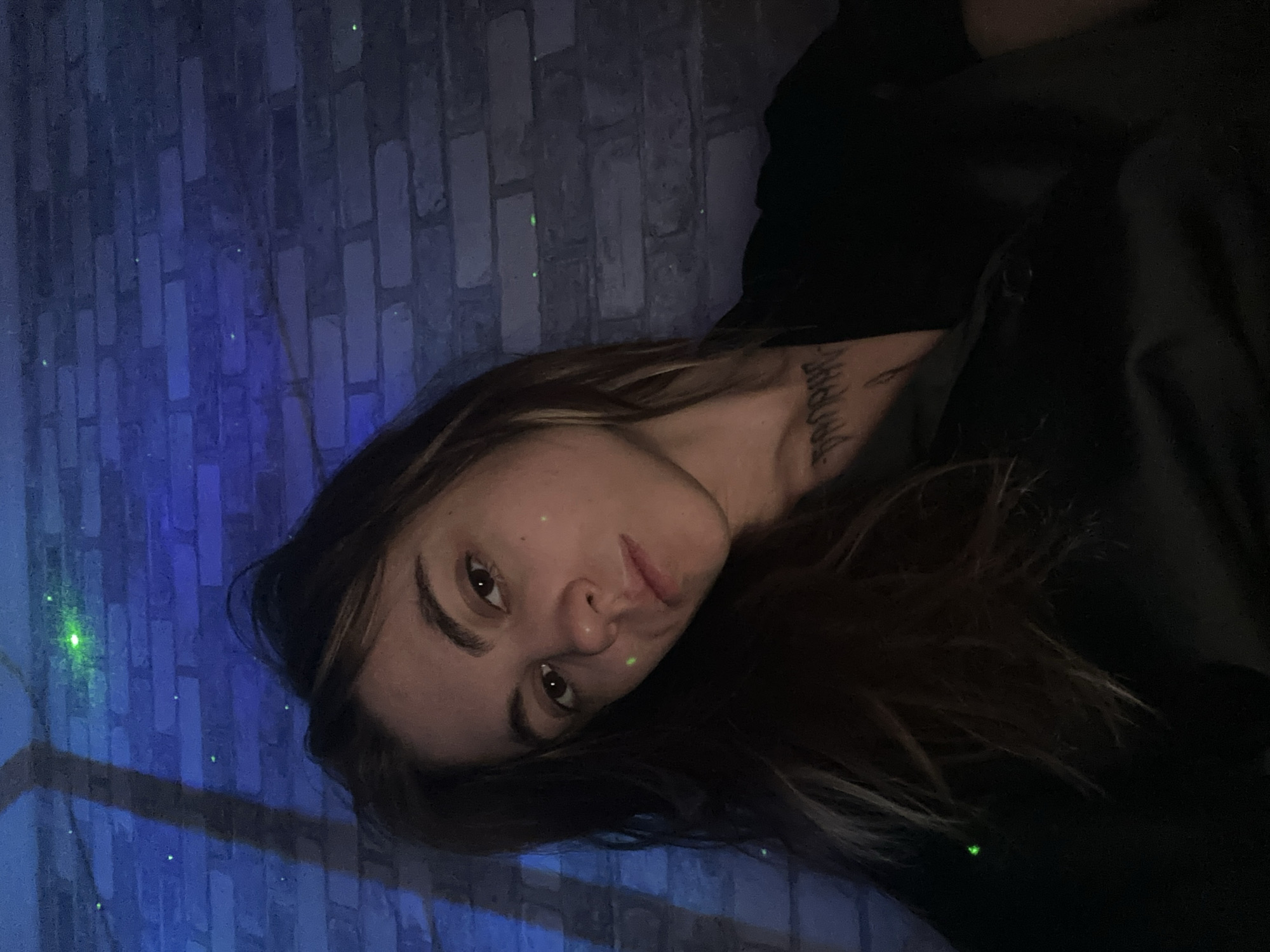 A woman is pictured in a selfie taken indoors against a brick wall. The wall is illuminated by blue and green lights, creating a dim and atmospheric setting. The woman has long, brown hair that falls over her shoulders and is wearing a dark top. A tattoo is visible on her chest. The image appears to have been taken in a dimly lit environment, possibly a club or a home decorated with special lighting. There is no indication of the specific location within Moscow, Russia.