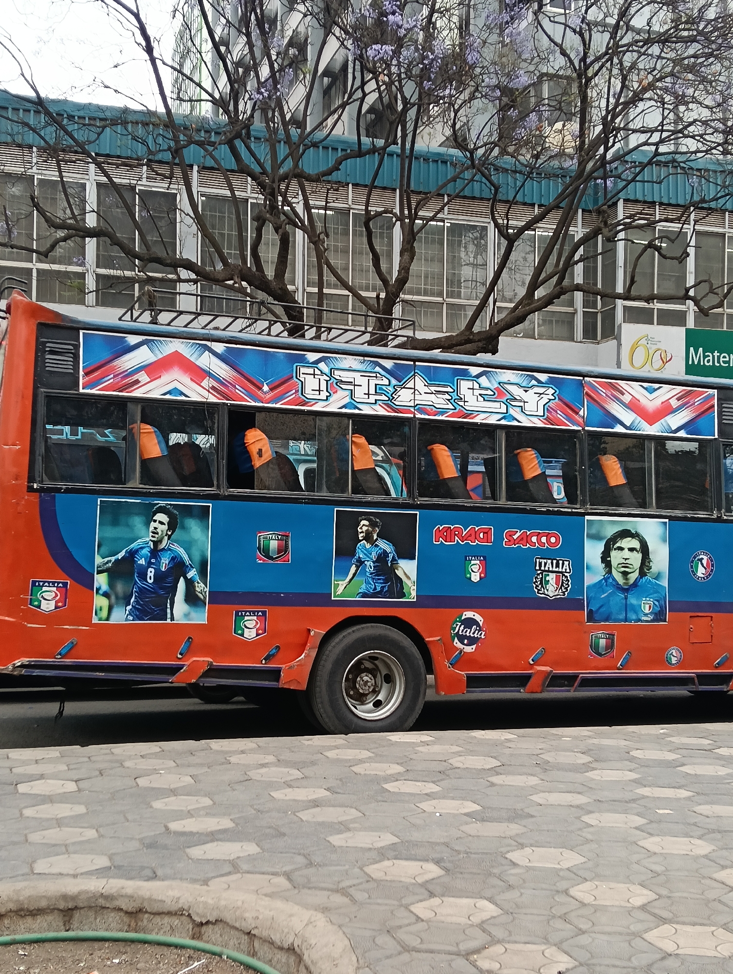 A side view of an orange and blue public transport bus, a matatu, is parked on a paved street in Nairobi, Kenya. The bus is decorated with images of Italian football players and the word "ITALY" in large lettering, along with Italian flags. The bus has tinted windows, and orange seat covers are visible inside. The bus is branded with "KIRADI SACCO". In the background, a building with large windows and a tree with purple flowers can be seen. The ground is paved with hexagonal tiles.
