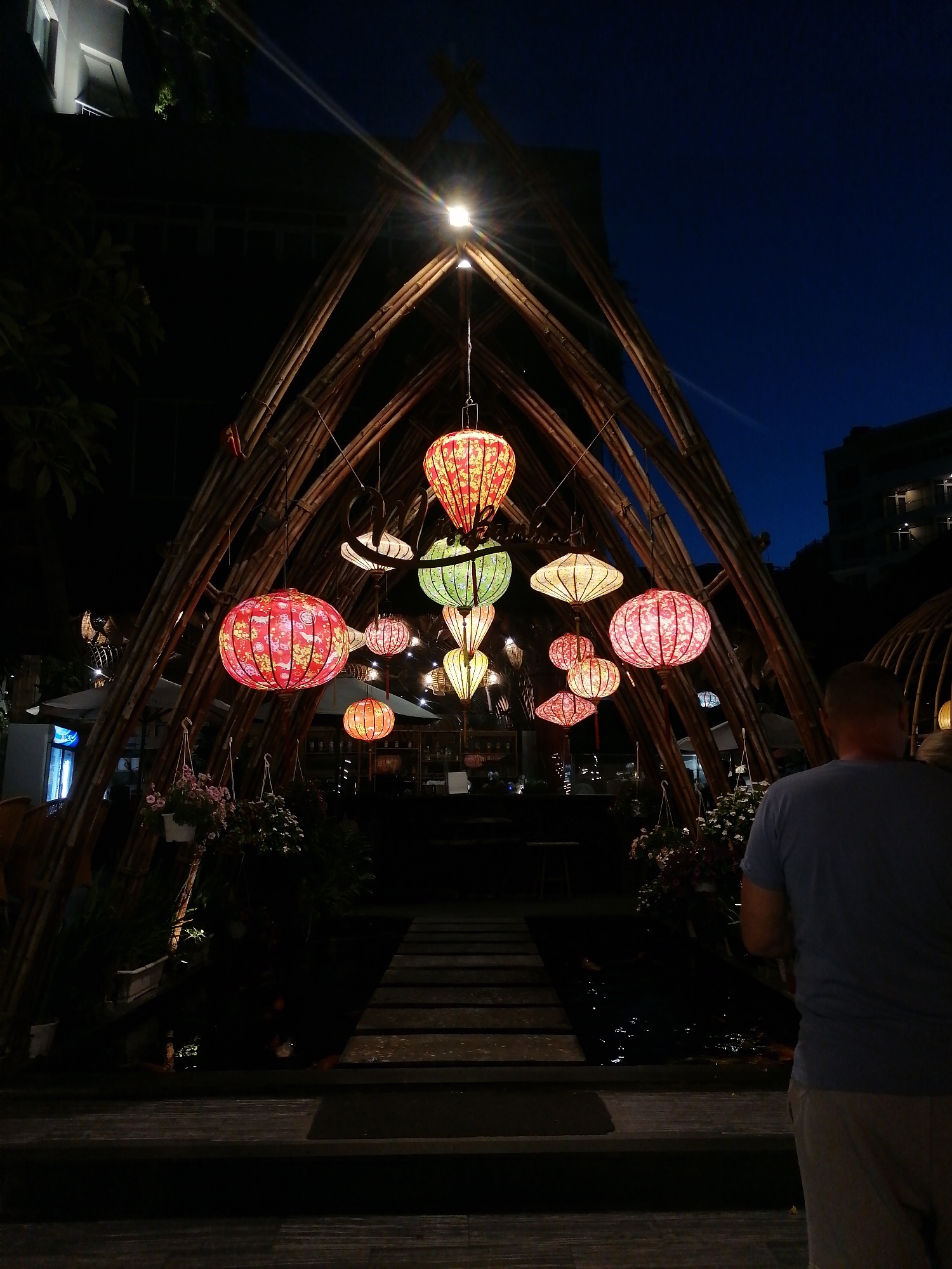 The image captures an outdoor setting in Nha Trang Ward, Vietnam, at night. A large, triangular bamboo structure stands prominently in the scene, adorned with multiple colorful lanterns hanging from its apex. The lanterns vary in size, shape, and color, casting a warm glow on the surrounding area. The path through the structure is lit by the lanterns, with flower boxes on the left and right of the path. In the foreground, a man in a blue t-shirt and khaki shorts is seen from the back, standing to the right side of the frame. Beyond the bamboo structure, the lights of other buildings are visible against the dark blue sky. The area is dimly lit, and the overall scene evokes a tranquil and festive atmosphere.