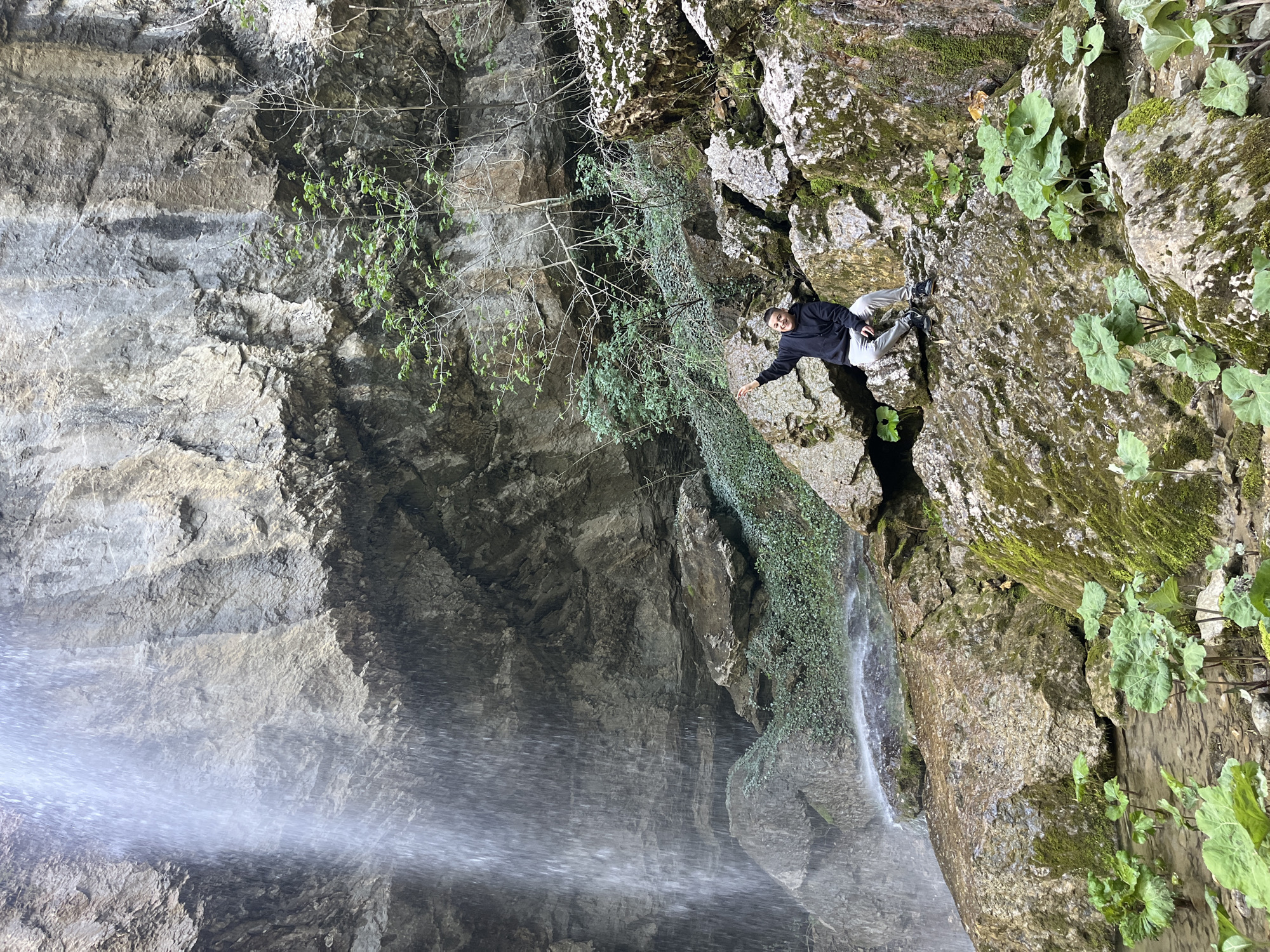 A vertical outdoor shot shows a man in a black jacket and light-colored pants standing on a mossy rock ledge near a waterfall. The waterfall cascades down the left side of the image. The rock face behind the man is covered in moss and some green plants. Some plants are visible on the rocks to the right of the man. The shot is taken in Veselinovo, Bulgaria.