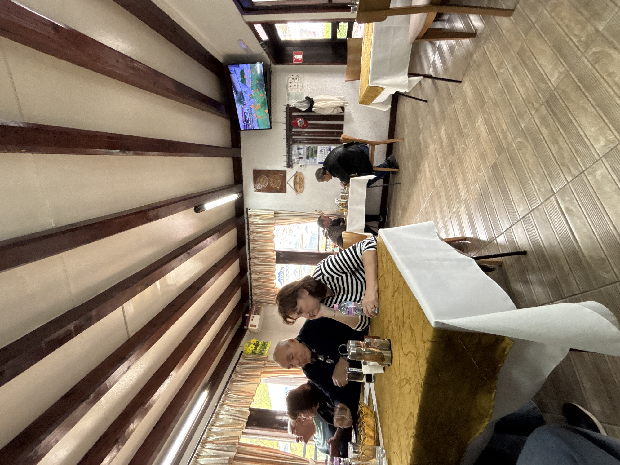 The image shows the interior of a restaurant, potentially in Zlatograd, Bulgaria. The restaurant has a rustic design, with wooden beams on the ceiling. A television screen is mounted on the wall displaying a program. Several people are seated at tables covered with white tablecloths, apparently eating. One woman wearing a black and white striped shirt is leaning on the table. Other people are facing the table. The floor is tiled. There are windows letting in natural light. The environment suggests a relaxed and casual dining atmosphere.
