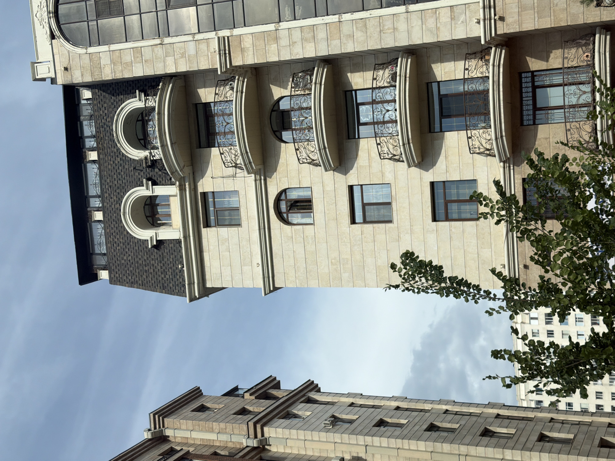 The image shows a view of several modern buildings in Astana, Kazakhstan, against a backdrop of a cloudy sky. On the right, a tall building with a beige stone facade features multiple balconies with ornate iron railings. The balconies have curved, projecting floors and are adorned with decorative metalwork. Above these balconies, there are arched windows, and the building has a dark, mansard-style roof with dormer windows. To the left, another building with a similar beige stone facade rises, showing a more uniform architectural style with rectangular windows. In the foreground, a leafy green tree partially obscures the lower portions of the buildings, its branches reaching upwards. In the distance, other buildings, including one with a white facade and numerous windows, are visible. The overall scene depicts an urban environment characterized by contemporary architecture.
