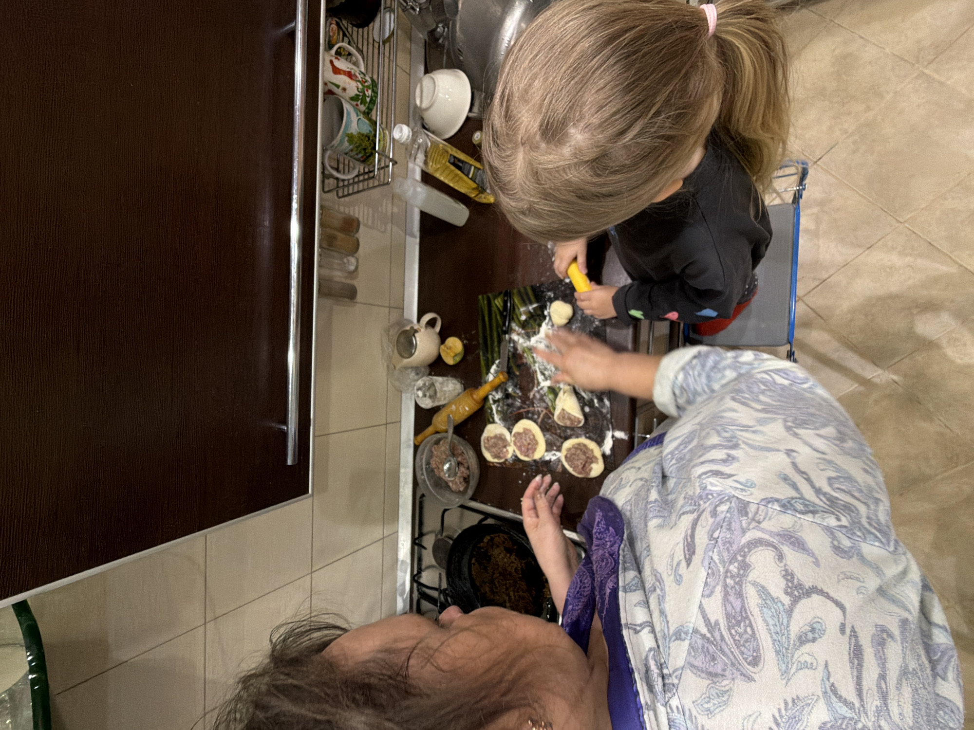 This top-down view captures an adult and a young child engaged in food preparation within a home kitchen in Kamyshin, Russia. The adult, seen from the back of their head with dark hair, is positioned in the foreground at the bottom. The child, with light brown hair tied in a ponytail, is seated on a stool or chair at a dark countertop, actively involved in the cooking.
The activity appears to be the making of dumplings, pelmeni, or vareniki. Bowls containing raw dough and minced meat filling are visible on the counter. The child holds a yellow tool, seemingly using it to press or shape a piece of dough, while an adult's hand is seen assisting with forming the dumplings. Several partially assembled dumplings, filled with meat, are arranged on the counter alongside a wooden rolling pin.
The background shows a cluttered but functional kitchen space with a sink, a dish drying rack holding mugs and bowls, and various bottles, including one of cooking oil. The floor is covered with light-colored square tiles. Dark wooden kitchen cabinets with a metal handle are visible along the left side. The scene conveys a collaborative and domestic cooking experience. No visible text is discernible in the image.