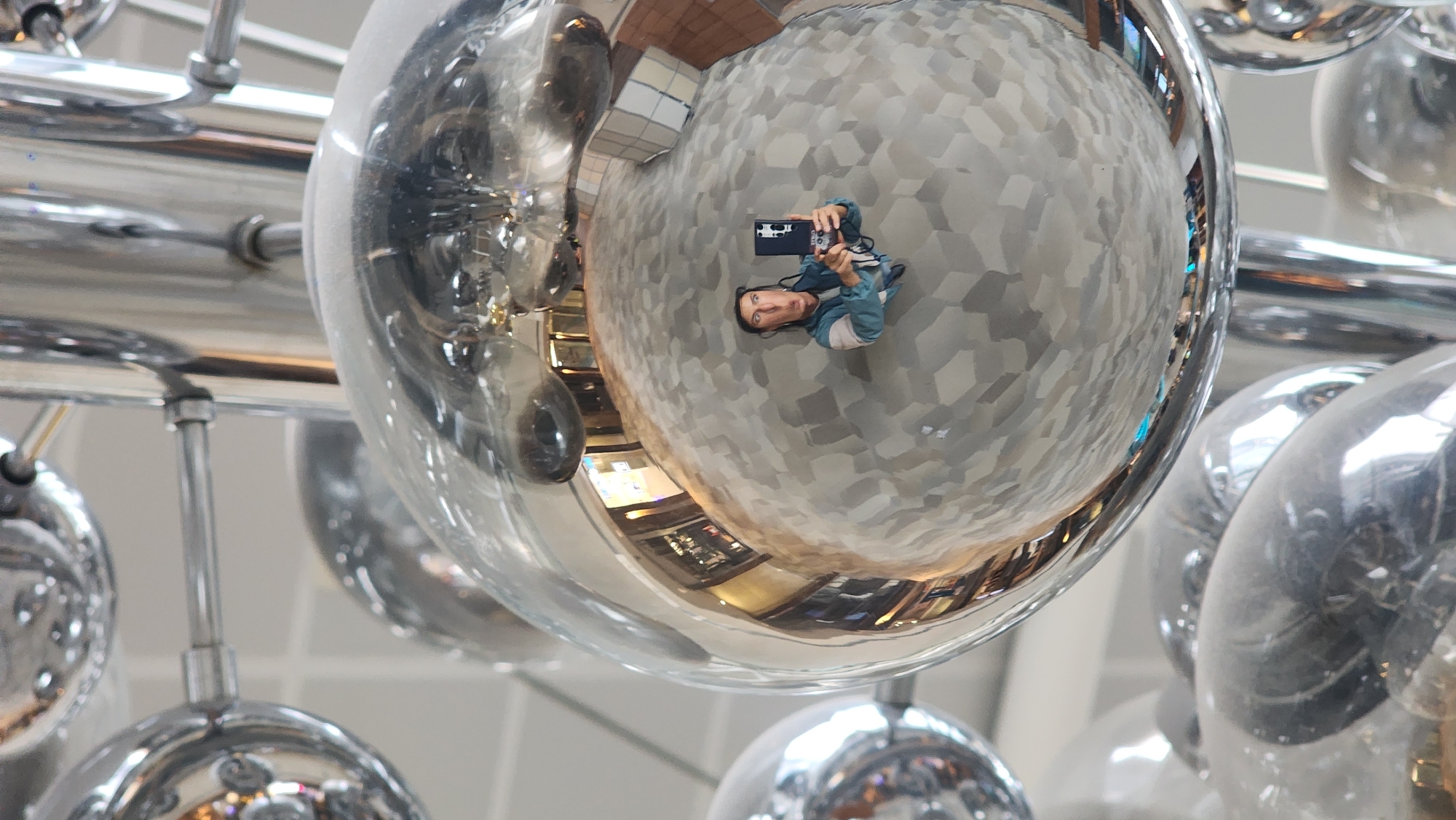 The image captures a reflective art installation of multiple metallic spheres, possibly a chandelier, in an interior space. A person is reflected in one of the larger spheres, holding a phone and taking a picture, indicating a selfie. The reflection shows a tiled floor and portions of a building interior, suggesting the artwork is located in a public space, potentially a shopping center or airport. The design of the tiled floor has a three-dimensional geometric pattern of gray cubes. The overall visual effect is distorted and fragmented due to the spherical reflections. Based on the art installation and architectural design of the interior, it is likely that this picture was taken in the city of Moscow, Russia.