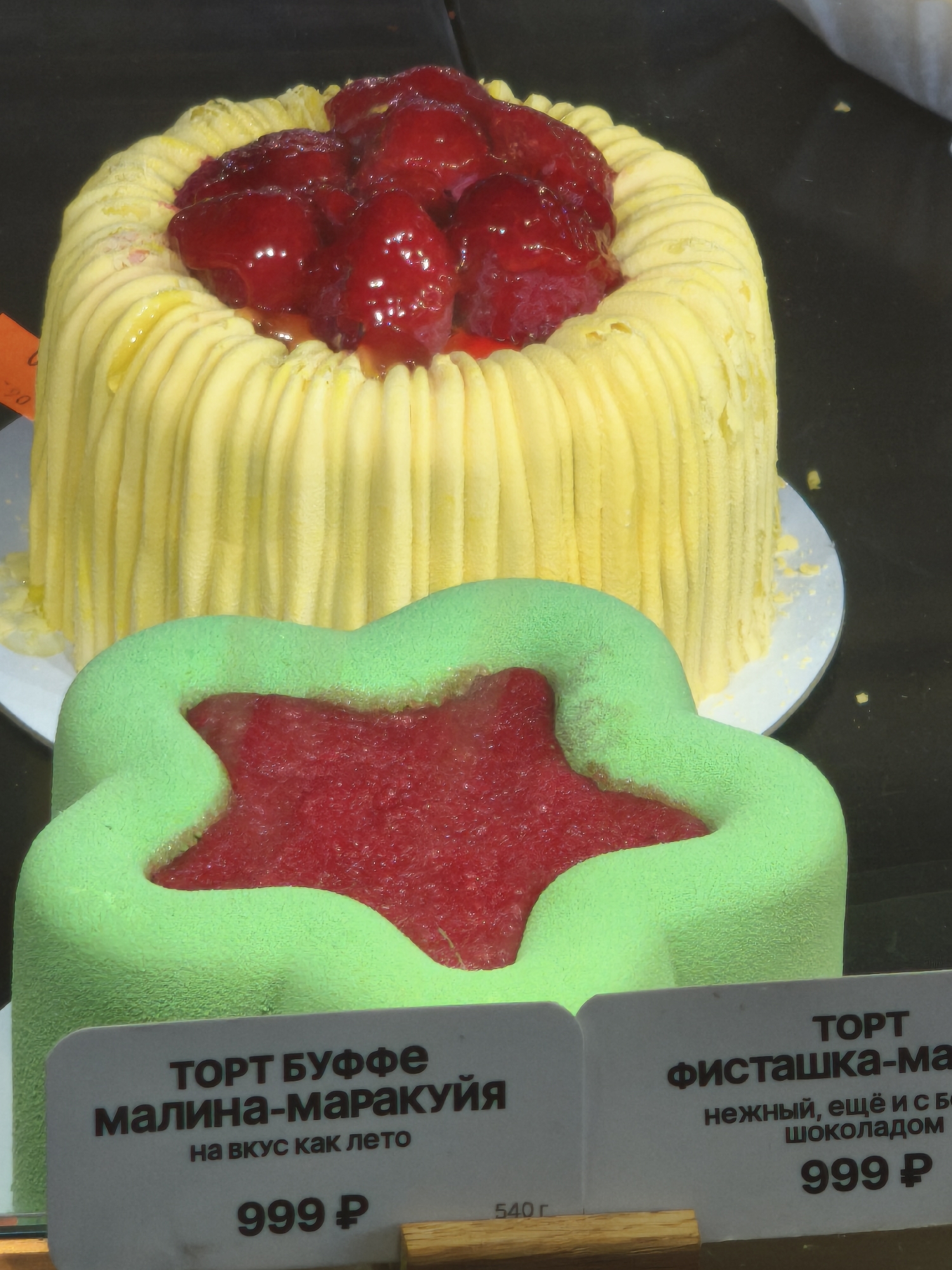Two cakes are displayed on a dark surface. The cake in the background is round with vertical yellow frosting and topped with red berries. The cake in the foreground is star-shaped with green frosting and a red filling. Two price tags are visible, one for the star-shaped cake at 999 ₽ and another partially visible price tag for the round cake, also showing 999 ₽. The scene appears to be a bakery or dessert display, possibly in Medovoye, Russia, given the currency and Cyrillic text on the price tags.