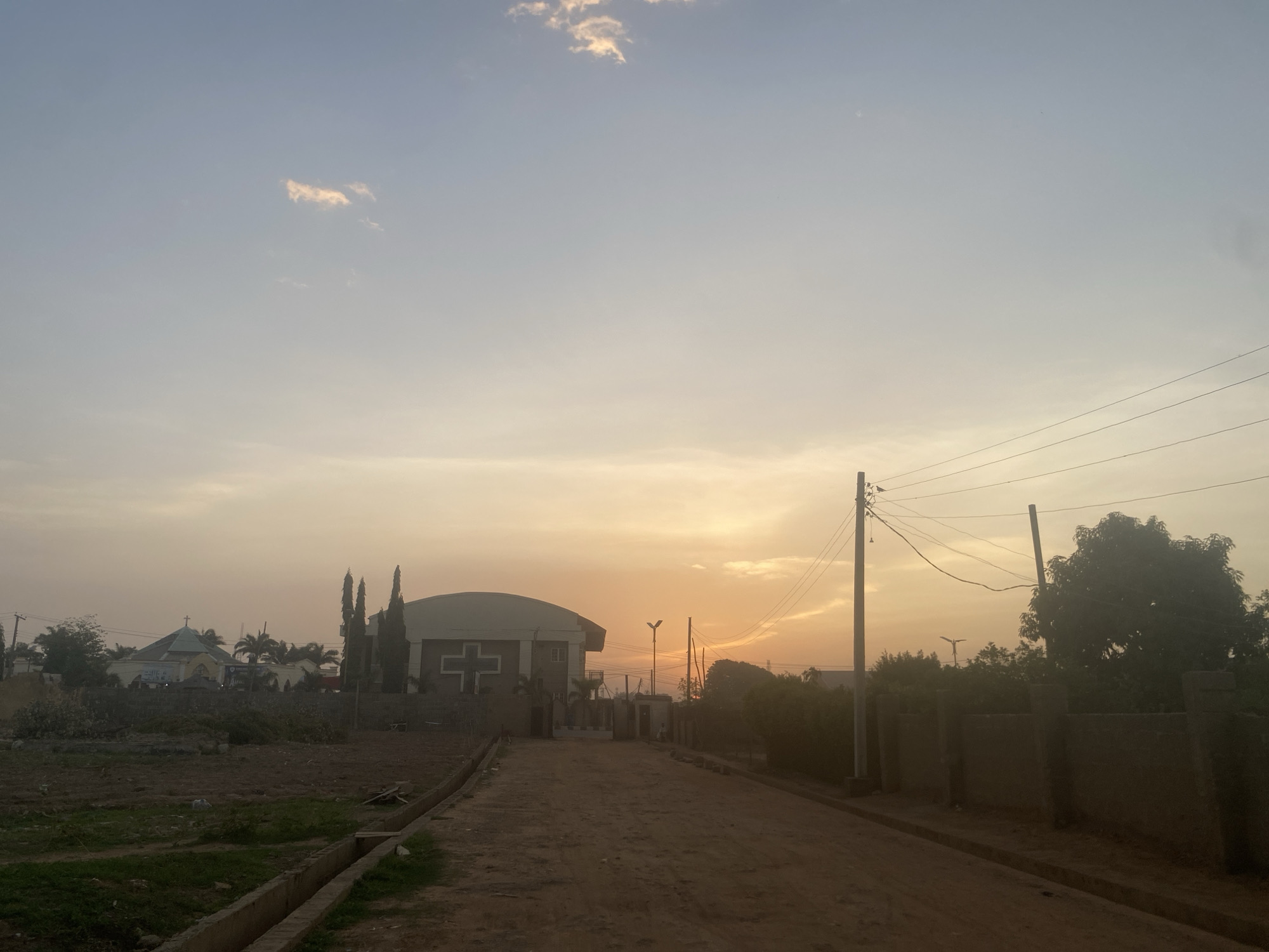 The image shows a dirt road in Barden Yana, Nigeria, at sunset or sunrise. On the left, there's a vacant lot with sparse vegetation and a church building visible in the background. Another church building, larger in size and sporting a cross on the facade, sits further down the road. The right side of the road is bordered by a wall with trees behind it and a utility pole with wires. The sky is bright with the colors of dawn or dusk.