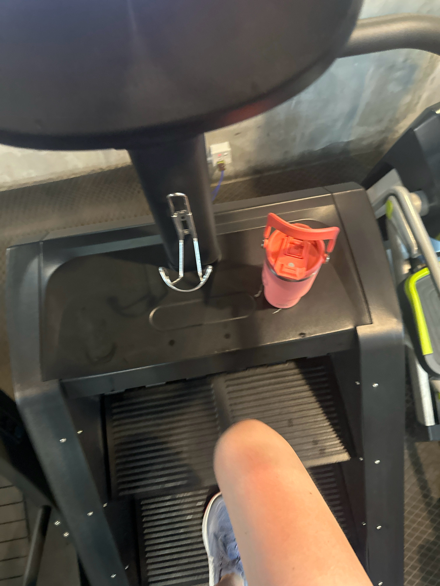 The image shows a close-up, high-angle view of a person's leg and foot on a treadmill. The treadmill deck is visible, as is a pink water bottle with a handle. The background appears to be a gym with a concrete wall and electrical outlets. The person is wearing a blue athletic shoe. The image does not provide any information about the location in the city of Lalitpur, Nepal.