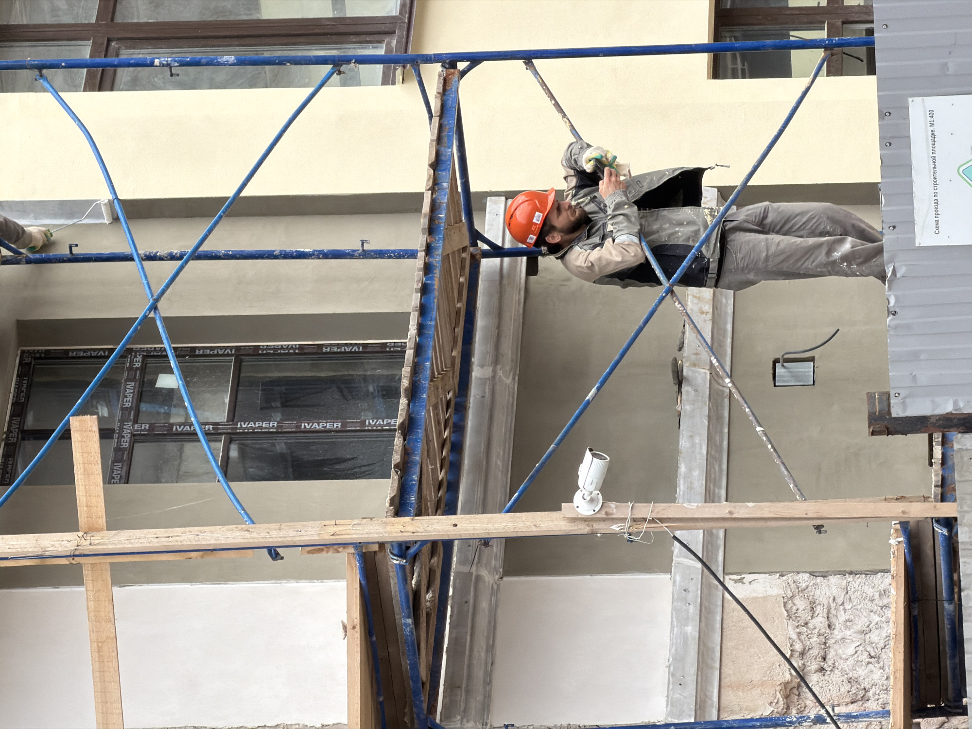 A male construction worker wearing an orange hard hat and a grey work jacket is standing on scaffolding outside a building. He is looking down at his phone, holding it in his right hand while his left hand, wearing a green glove, rests on the scaffolding railing. The worker is positioned on the second level of the scaffolding. The building appears to be under renovation, with exposed plaster and scaffolding. A security camera is mounted on a wooden pole to the left of the worker. The scene is outdoors with overcast lighting. The building's facade is painted a light beige color, and there are dark-framed windows visible. The image is a medium shot focused on the worker and the immediate surroundings of the scaffolding. There is a sign with Russian text on a metal fence in the foreground.