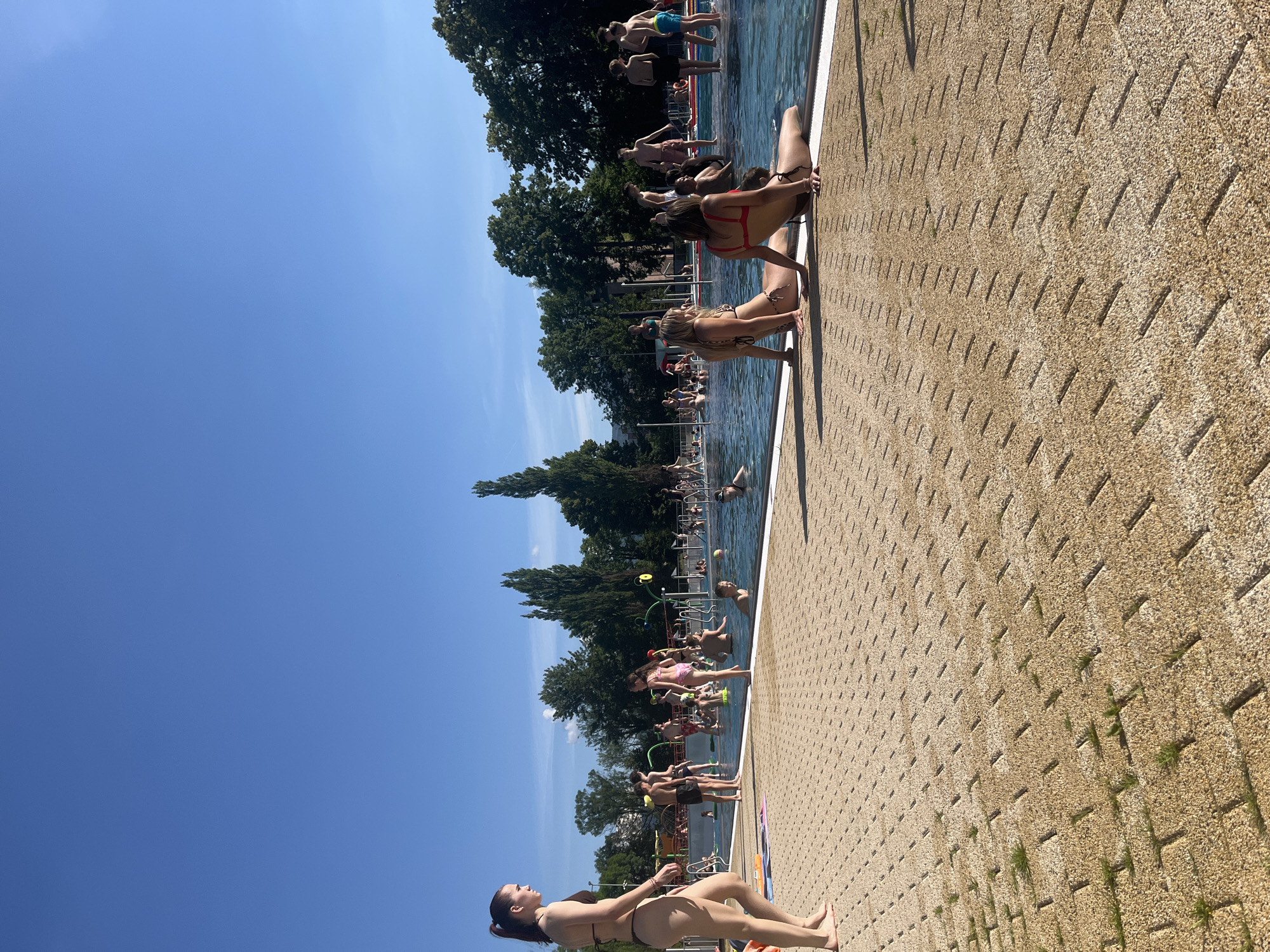 A full shot captures a lively outdoor swimming pool scene on a sunny day. The foreground features a tan, textured pavement with sparse patches of green grass growing between the paving stones. In the midground, a woman in a dark bikini walks towards the left, her back to the camera. Further into the pool, numerous people are engaged in various water activities. Some are swimming, others are standing or sitting in the water, and a few are interacting with water features, suggesting a family-friendly recreational area. The pool is surrounded by a white edge. The background is filled with lush green trees and a clear blue sky.
