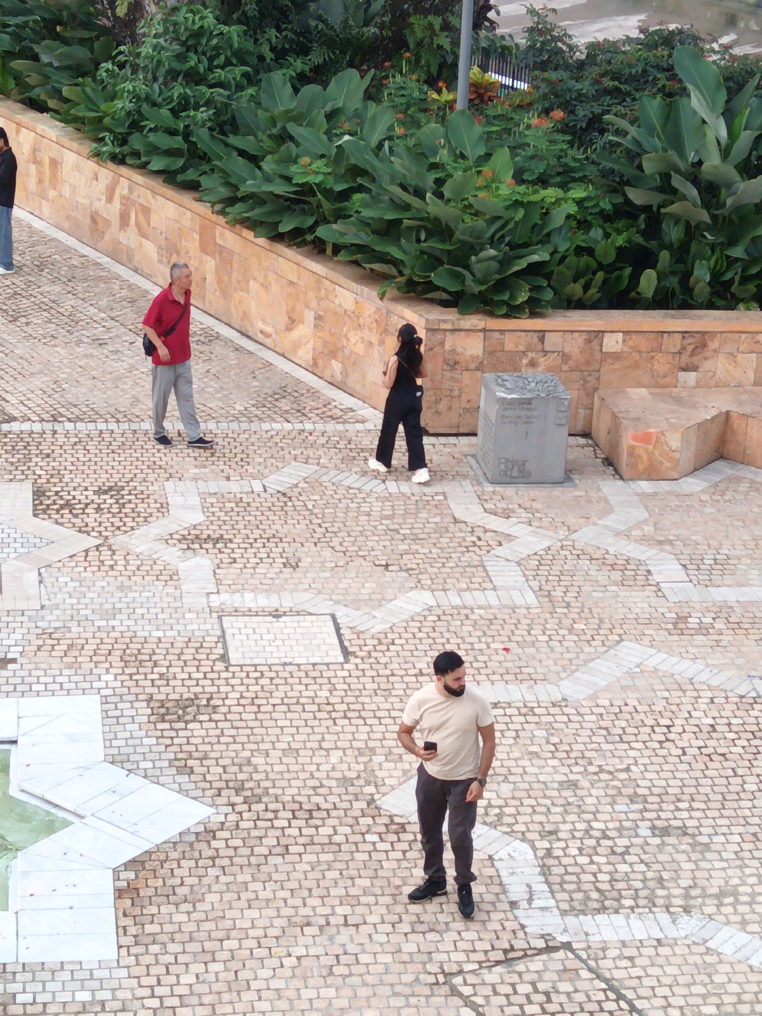 The image depicts an outdoor scene with people walking on a paved area. The paving consists of small, light-colored bricks arranged in a pattern, interspersed with larger white geometric shapes.
In the background, there is a terraced planter with lush green foliage and a textured stone wall. Further back, there appears to be more vegetation and possibly some fencing.
There are three individuals visible in the image:
1. A man wearing a red shirt and grey pants, walking towards the right. He has a dark bag slung over his shoulder.
2. A woman wearing a black sleeveless top and black pants, walking towards the left. She is wearing white shoes.
3. A man wearing a light-colored t-shirt and dark pants, standing and looking down at his phone. He has a beard and is wearing black shoes.
In the mid-ground, there is a large, grey, cubic sculpture with some text on it, placed on the paved area. To the right of the sculpture, there is a large, light-brown stone structure.
The overall environment suggests a public space, possibly a park or plaza, within a city. The specific location in Kuala Lumpur, Malaysia, is not identifiable from the visual cues in the image.