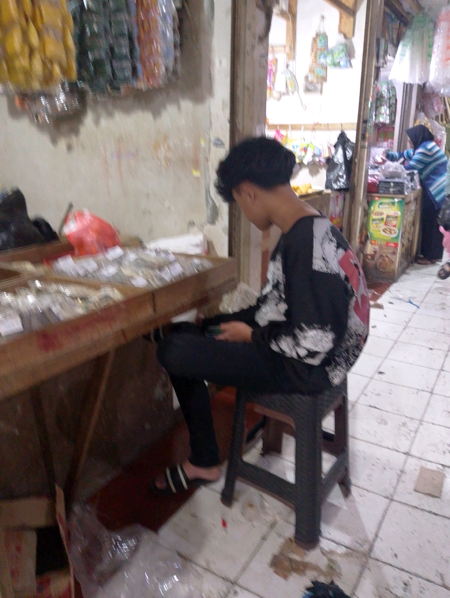 A medium shot shows a young male sitting on a dark plastic stool in front of a wooden display table. He is wearing a black long-sleeved shirt with a white and red abstract graphic print and black pants. His head is turned to the right, and his gaze is directed downwards. The wooden table in front of him is filled with small items, possibly accessories, arranged in compartments. A red plastic bag is visible on the table.
The environment appears to be an outdoor market stall in Lembang, Indonesia. Behind the table, a doorway leads into a dimly lit interior of a shop. The shop's interior displays various goods, including what appear to be packaged snacks or small items. To the right of the frame, a woman wearing a blue and white striped hijab and a blue striped top is interacting with a counter. The ground is tiled with white squares. The wall behind the young male is a rough, concrete texture. The overall lighting suggests it is daytime.