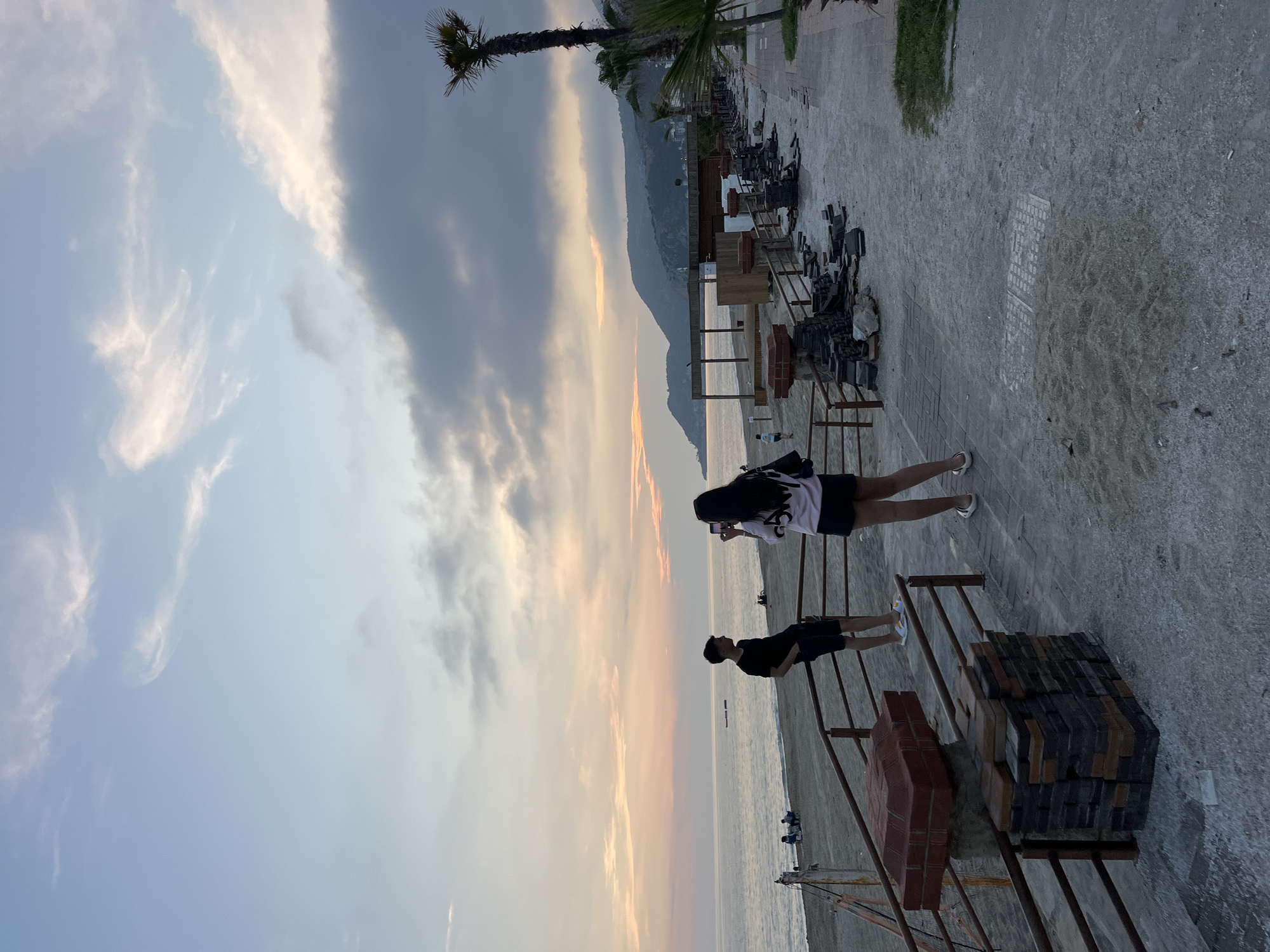 The image captures a picturesque coastal scene, likely a promenade or pier, in Alanya, Turkey, during either sunrise or sunset. Two individuals stand prominently in the mid-ground, facing the sea. The person on the right has long dark hair and wears a white top, dark shorts, and white sneakers, holding what appears to be a phone, potentially taking a picture. To their left, another person in a dark t-shirt, dark shorts, and white sneakers also looks out towards the water.
In the immediate foreground and to the right, stacks of paving stones or bricks are arranged on metal frames, suggesting ongoing work or temporary installations. A tall palm tree with a slender trunk is visible on the far left, adding a tropical element to the landscape.
The calm sea stretches towards the horizon, where a faint, elongated object is visible. Along the coastline, a series of structures, possibly beach bars or small buildings, are dotted along the shore, backed by majestic mountains that recede into the distance.
The sky is a dominant feature, displaying a beautiful gradient of colors characteristic of dawn or dusk. Soft blues near the top transition into warm oranges and pinks closer to the horizon, with wispy white clouds scattered across the lighter areas and darker, heavier clouds gathered towards the upper left. The lighting is soft and golden, creating a peaceful and serene atmosphere. A faint, rectangular grid pattern is visible on the concrete ground.