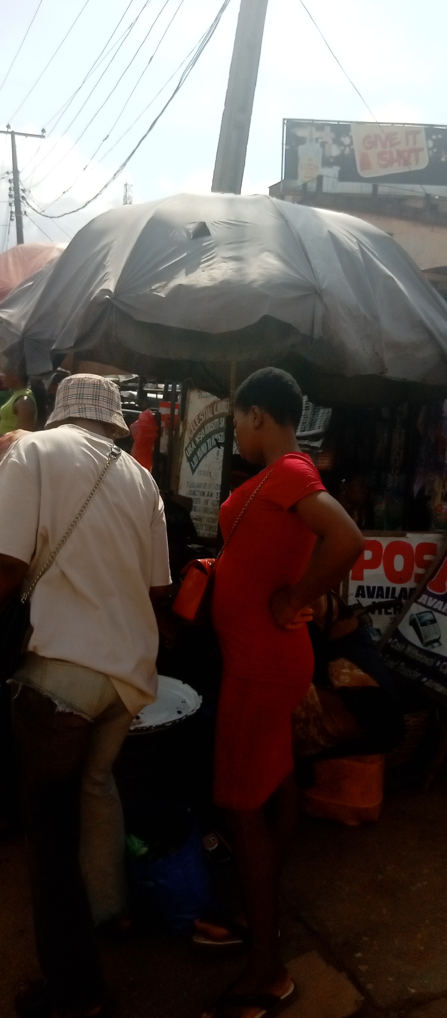 The image shows a street scene at Nkpor, Nigeria. Two individuals are present: one is facing away from the camera wearing a white shirt, khaki shorts, and a patterned bucket hat; the other person, dressed in a red dress, faces right with their hand on their hip. They appear to be standing near a market stall covered by a gray tarp. In the background, there is a billboard with the text "GIVE IT A SHOT" and a sign that reads "CELESTIAL CH" and "POS AVAILABLE".
