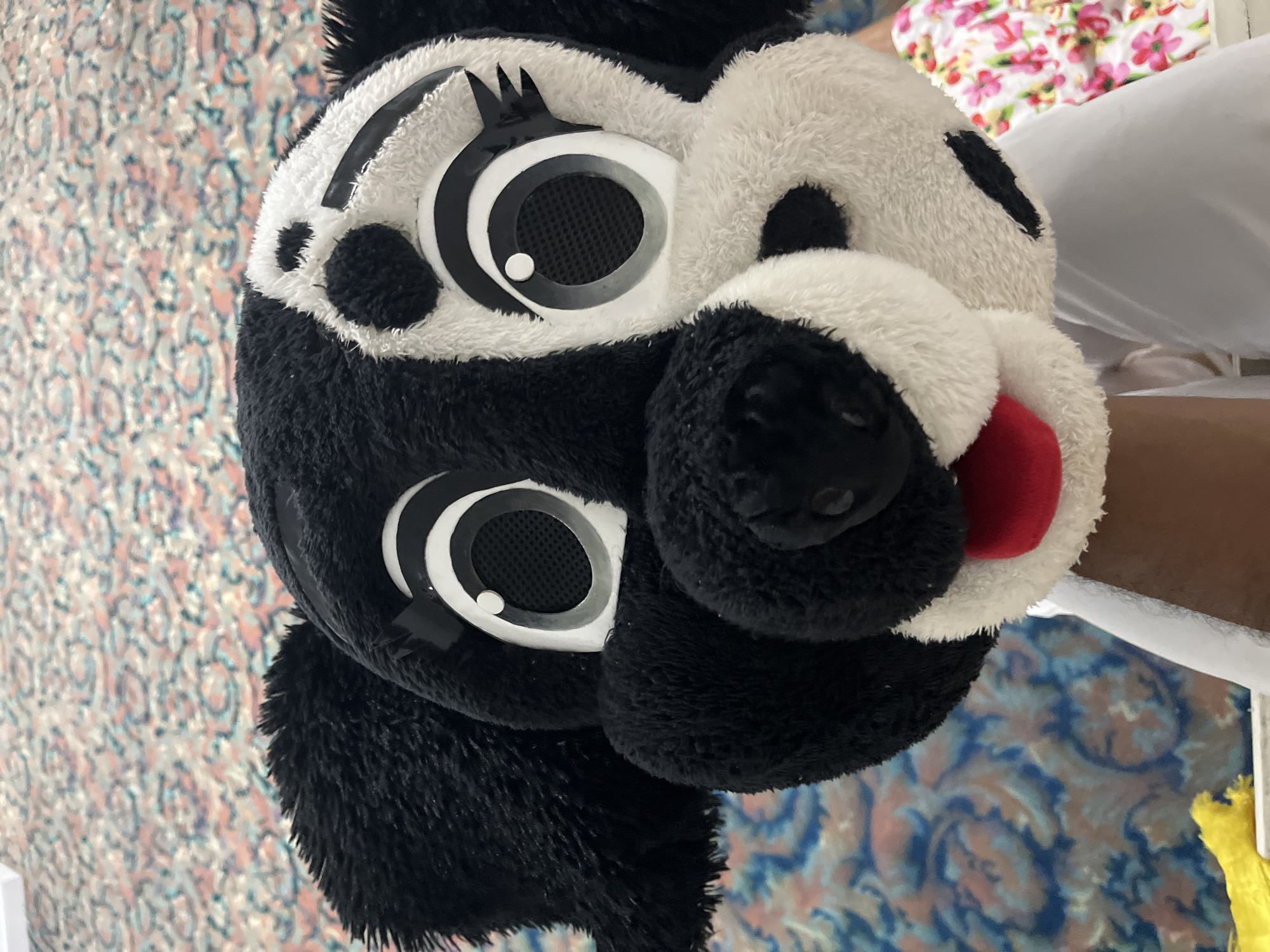 The image presents a close-up view of a large, black and white plush animal head, likely a panda, which appears to be either a costume head or a substantial stuffed toy. The head is made of soft, shaggy faux fur. Its most striking features are the oversized, stylized eyes, which are predominantly white with prominent black concentric circles and a mesh-like dark center, possibly allowing for vision if worn as a costume. A small white dot on each eye simulates a light reflection. Above the eyes, black, pointed shapes suggest stylized eyelashes or brow markings. A fluffy black snout with a distinct red, tongue-like feature peeking out from below it adds to the character's expression.
The background is an indoor setting with various soft textiles. To the left, a fabric with a faded, intricate pattern in shades of blue, pink, and off-white is visible, resembling a blanket or curtain. To the right, at the top, a section of white fabric with a cheerful floral pattern of pink, yellow, and green flowers is seen. Below this, a significant portion of plain white fabric, consistent with bedding, is present, partially obscuring a light brown surface, which could be skin or furniture. A thin white bar or rail and a small, fuzzy yellow object are also visible in the lower right corner. No specific activity is taking place, and there is no visible text in the image. The scene suggests a casual, domestic environment.