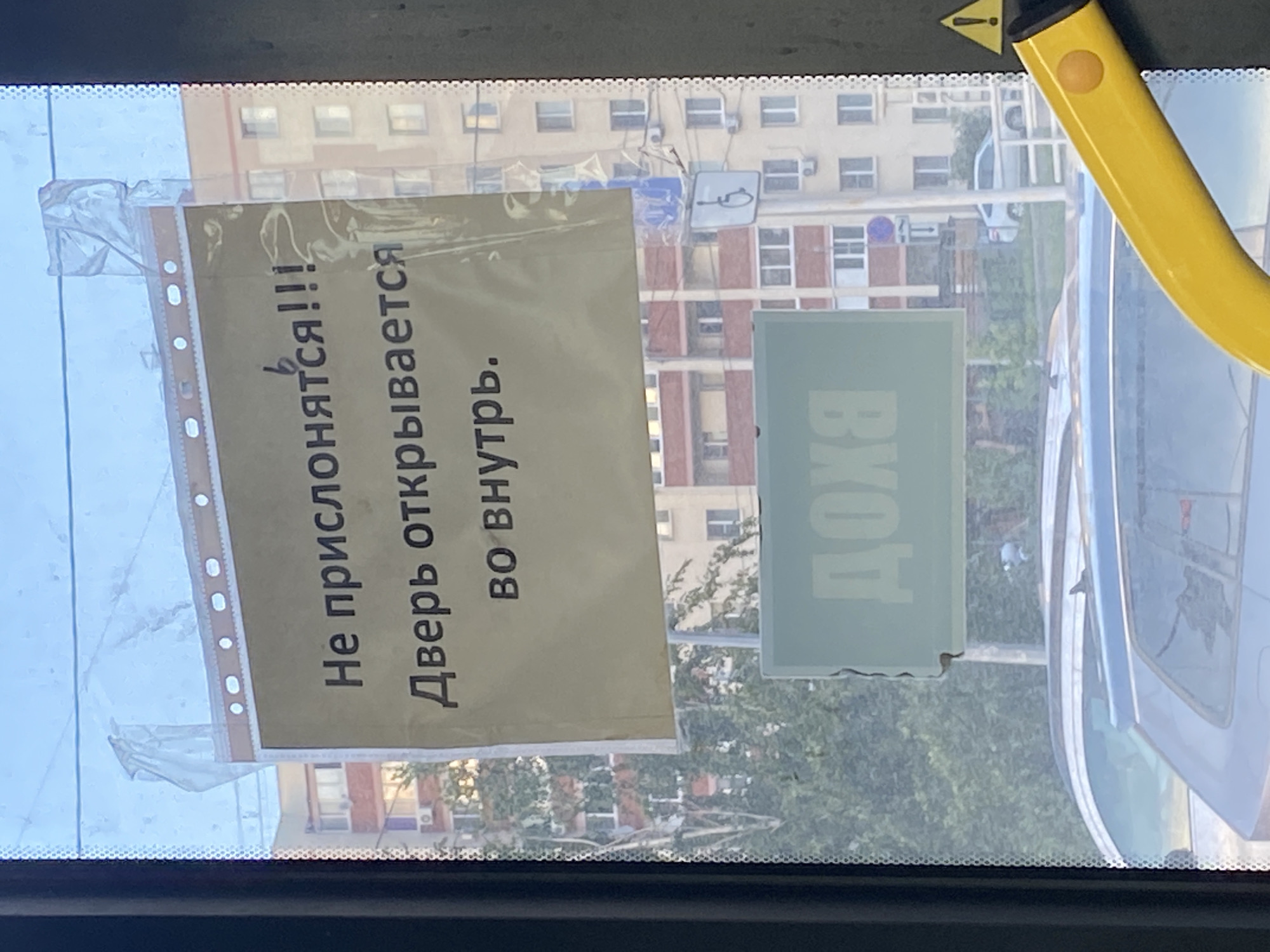 The image captures a view from inside a vehicle, likely a bus, looking out through the windshield. The scene is set during the daytime, as indicated by the bright sky and visible sunlight reflecting off surfaces.
The primary subjects are two signs attached to the interior of the windshield. The larger sign, oriented vertically, is a light brown paper in a clear plastic sleeve. It contains text in Russian. The text reads, "Не прислонятся!!!" which translates to "Do not lean!!!" and "Дверь открывается во внутрь." which translates to "The door opens inward." The second sign, a lighter green rectangle, also displays Russian text in a lighter shade, reading "ВХОД," meaning "Entrance."
The background visible through the windshield shows an urban environment with multi-story apartment buildings. Some buildings have visible architectural details like balconies and windows. Trees with green foliage are also present, suggesting a warm season. The weather appears clear and sunny.
Other visible objects include a yellow grab handle, common in public transportation, and a yellow triangular warning sticker with a black exclamation mark at the top of the windshield. Reflections on the glass obscure some details of the exterior scene and the vehicle's interior. The overall impression is a candid snapshot of everyday life within a public transport setting.