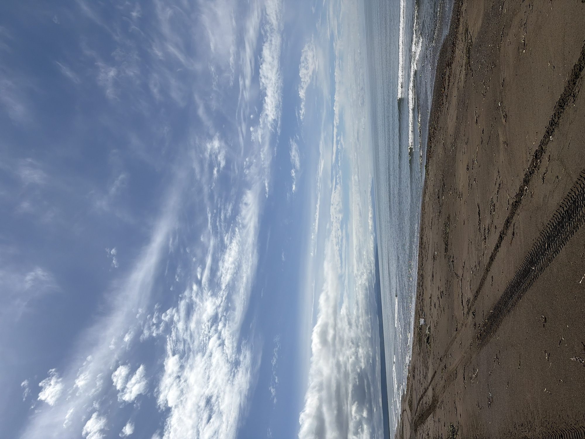 A wide shot captures a tranquil beach scene under a dynamic sky. The foreground is dominated by dark, wet sand marked by prominent tire tracks, indicating recent vehicular activity. Scattered debris, possibly seaweed and driftwood, lies on the sand. The middle ground features a calm sea with gentle waves lapping the shore. In the distance, a thin line of land is visible, with a hazy mountain range forming the horizon under a vast expanse of blue sky. Wispy white clouds streak across the sky, some appearing more solid and dramatic closer to the horizon. The overall impression is one of natural beauty and solitude. The location is identified as Ozerskoe, Russia.