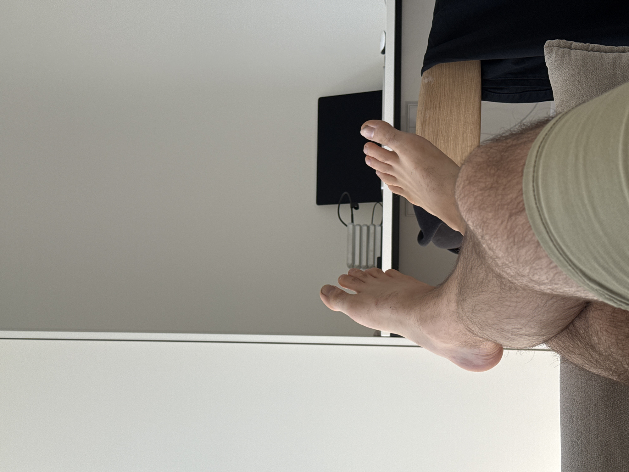 The image, when correctly oriented, captures a close-up view of a person's legs and feet in an indoor setting. The majority of the background consists of a plain, light-colored wall. On the right side of the background, a dark, rectangular screen or monitor is mounted on the wall, with cables visible beneath it.
In the foreground, a person's legs are prominently featured. The right leg, which is hairy, is bent at the knee, with the lower leg and foot extended towards the left. The foot is bare, showing toenails. A part of the person's left foot is also visible further back in the frame. To the immediate right of the person's legs, a section of a grey, upholstered sofa or chair with a matching cushion is visible. A light-colored wooden surface, possibly a desk or shelf, is situated behind the legs, with a dark garment draped over it.
The lighting is bright and even, indicating it is daytime. The overall ambiance suggests a casual and relaxed atmosphere, with the person likely lounging or resting. There are no specific activities depicted beyond this state of repose. No visible text is present in the image. The setting appears to be a modern interior space, consistent with a residence or hotel room in Kitsjanovo, Russia.