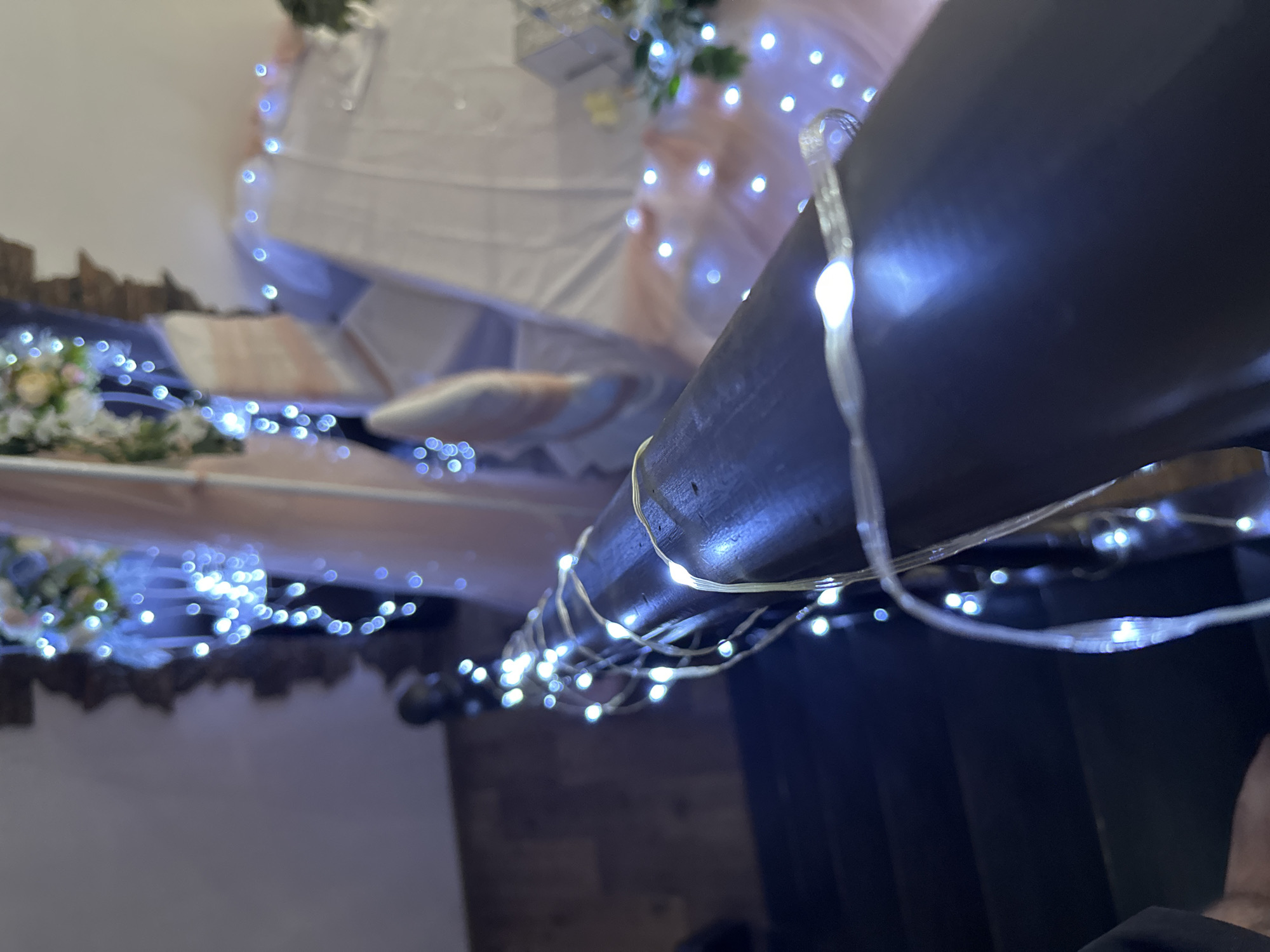 The image depicts a scene at an indoor event, likely a wedding reception, location Unknown, Unknown. A white tablecloth-covered table is adorned with floral arrangements and fairy lights. A dark-colored post is wrapped with fairy lights, creating a foreground element. The background is dimly lit, with dark vertical panels. A person is partially visible at the bottom right.