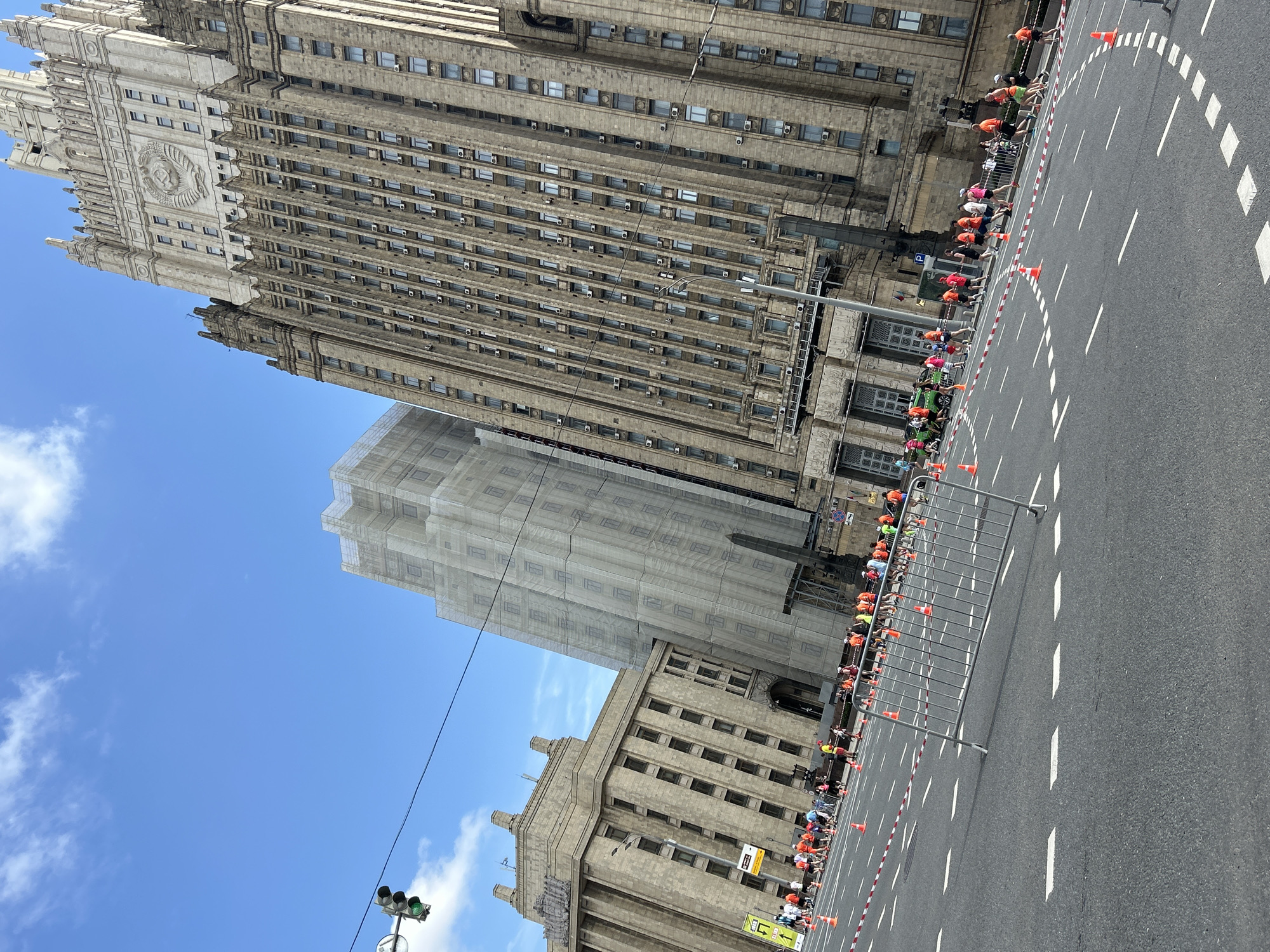 The image depicts a cityscape in an unknown location. A tall, light brown stone building with numerous windows and intricate architectural details dominates the frame. A second building, partially obscured by scaffolding or protective netting, stands alongside it.
A street runs along the right side of the image, demarcated by white lines and orange cones. A metal barrier lines the edge of the street, separating it from a gathering of people dressed in colorful athletic wear. They appear to be lined up, possibly participating in an event or race.
In the lower left corner, a traffic light is visible against a bright blue sky with scattered clouds. Power lines stretch across the sky, adding to the urban setting.
