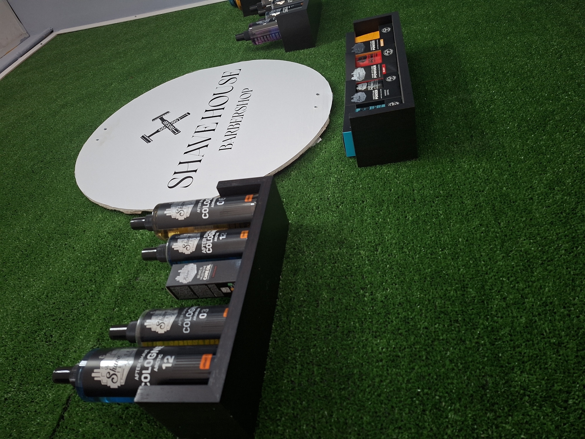 The scene depicts a studio shot of "Shave House Barbershop" products, possibly for advertising. The objects include a circular sign with the words "Shave House Barbershop" printed on it, several black shelves filled with aftershave cologne bottles labeled "Arctic 12" and "Arabia 03", and boxes of hair styling powder. The environment appears to be a studio setup with green artificial turf as the backdrop. There are no people visible in the image. The absence of real-world contextual cues prevents a precise location in Galabovo, Bulgaria, or specific actions or interactions.