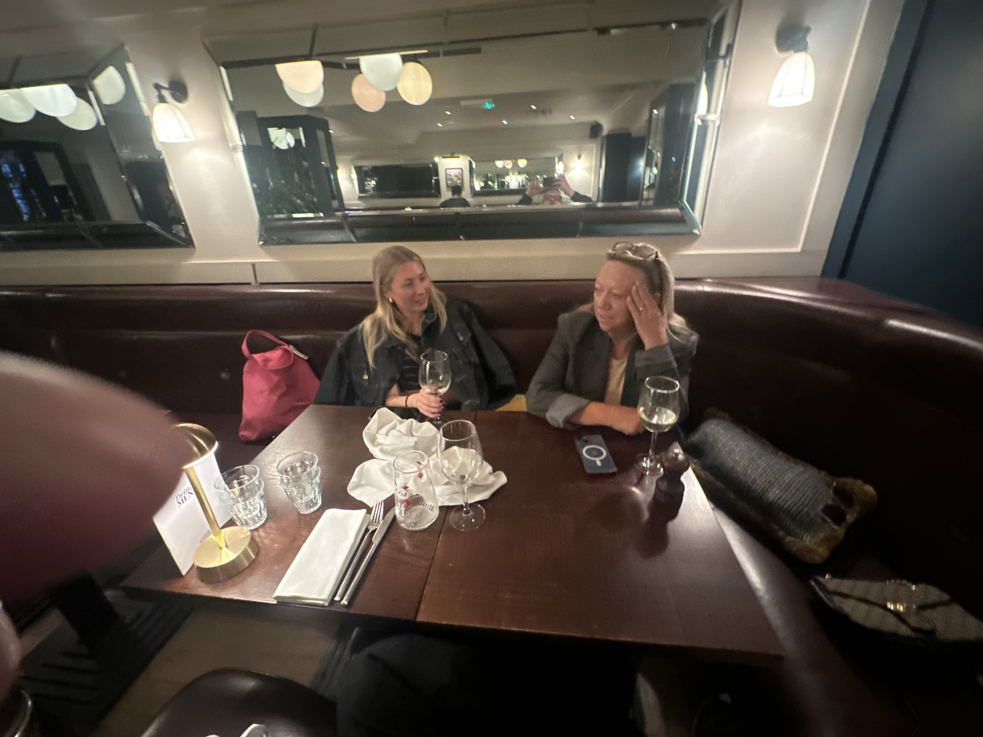 Two women are seated at a dark wood table in what appears to be a restaurant or bar. The location is likely in St Albans, United Kingdom. A cushioned booth wraps around them. A large mirror adorns the wall behind them, reflecting the interior of the establishment. The woman on the left has blonde hair and is holding a wine glass. The woman on the right has blonde hair pulled back and is wearing glasses; her hand is raised to her head. The table is set with glasses, silverware, napkins, and menus. A pink bag rests on the booth next to the woman on the left. A cell phone is on the table next to the woman on the right. Additional bags are on the booth seat, to the right. Artificial lighting fixtures are visible on the walls and ceiling.