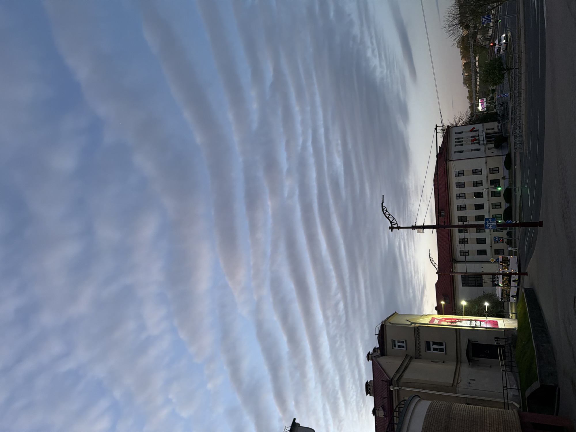 This image captures an urban street scene at what appears to be dusk or dawn, distinguished by a striking sky. The most prominent feature is the sky, filled with long, parallel, wave-like cloud formations, likely atmospheric gravity waves or Kelvin-Helmholtz instability clouds, stretching across the frame from left to right. These clouds display varying shades of gray and white against a light blue sky, creating a visually dramatic effect.
On the right side of the photograph, a row of buildings lines a street. A large, multi-story building with a light-colored facade, a prominent red-tiled roof, and numerous dark-framed windows is central to the urban element. Below its windows are visible flagpole mounts, some displaying red and white elements that could be flags. Adjacent to it, a smaller building with a similar architectural style has a large, illuminated banner or sign on its side, though the text is illegible. Further down the street, another building with a red roof is visible.
The street itself features streetlights, traffic signals (one showing a green light), and various road signs. In a cropped view, a white bus is seen driving on the road. Patches of green grass and some trees, mostly bare or with sparse foliage, are visible along the street and near the buildings. The overall lighting is soft and muted, with streetlights already active, contributing to the twilight atmosphere.
The scene is set in Hrodna, Belarus. No specific activities are ongoing; the image presents a static view of the city under a remarkable sky. No text is clearly readable on any signs, banners, or vehicles within the provided media.