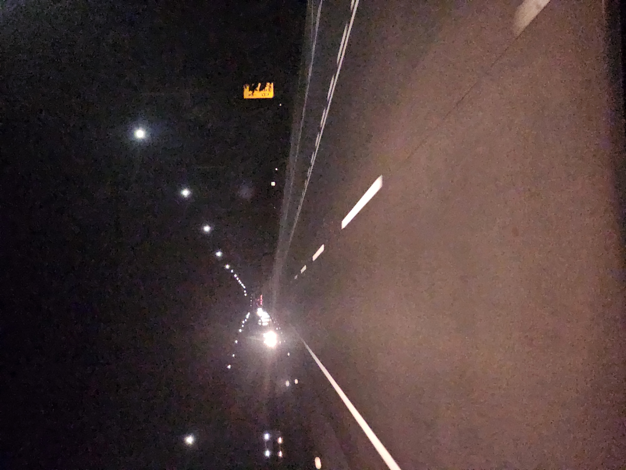 The image captures a nighttime view of a highway, likely in Sangju-si, South Korea, characterized by a dark sky illuminated by streetlights and vehicle headlights. The road, paved with asphalt, has distinct white lane markings, a solid white line on the left and dashed white lines indicating traffic lanes. Several vehicles are visible: on the left, headlights suggest a vehicle approaching, while further ahead, a line of headlights indicates a procession of cars. On the right side of the highway, a yellow rectangular sign with black silhouettes of deer is illuminated, warning of wildlife crossing. The overall impression is a well-lit highway at night, with traffic moving, and a reminder of the potential presence of animals.