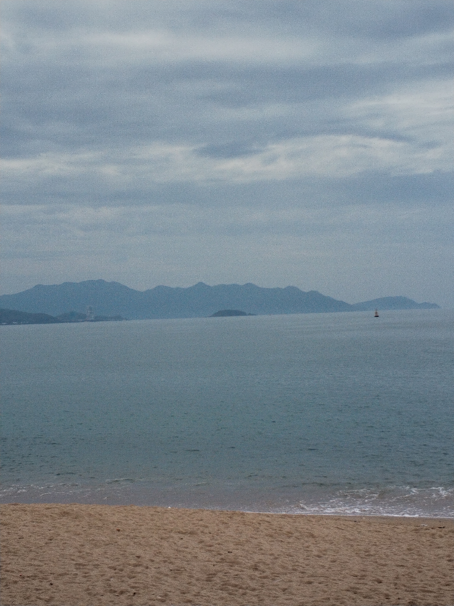 The image captures a tranquil coastal landscape, likely in Nha Trang Ward, Vietnam, under an overcast sky. In the foreground, a stretch of light brown sand forms a beach, extending across the bottom third of the frame. Gentle waves with white foam break softly where the sand meets the calm, blue-grey ocean.
Across the expansive body of water, a long range of hazy, blue-grey mountains rises in the distant background, forming a continuous silhouette against the sky. Near the base of these mountains, slightly to the left of the frame's center, a faint, tall structure—possibly a building or a tower—is visible. Further right in the mid-distance of the ocean, a small, distinct red and white buoy floats, a solitary object in the vast seascape.
The sky is entirely covered by soft, diffused light filtering through uniform light grey clouds, indicating an overcast daytime. The muted color palette of blues, grays, and earthy browns contributes to a calm and somewhat melancholic atmosphere. No people or specific activities are depicted. The overall image has a grainy texture. No visible text is present.