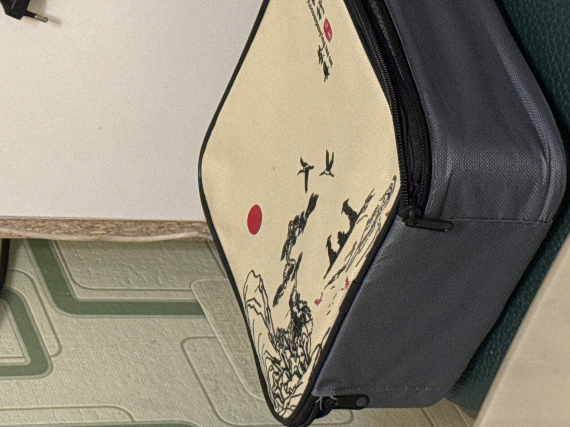 This media file presents a close-up, rotated view of a soft-sided carrying case with a distinctive design. The main subject is this case, which has an off-white fabric panel on the front, adorned with an East Asian-inspired ink painting. This design features a vibrant red sun, two black birds in flight, and a stylized landscape of mountains and foliage rendered in black brushstrokes.
The case itself is predominantly dark grey fabric, with visible stitching and a black zipper closure that runs along one of its edges, suggesting it opens to reveal an interior compartment. A small zipper pull tab is discernible at the bottom of the case.
In the top left corner of the image, part of a black electrical plug with two metal prongs is visible against a plain light-colored surface, possibly a wall. The background also includes a patterned surface with geometric lines in shades of light green and beige, resembling wallpaper or floor tiling. Additionally, a rough-edged wooden surface, possibly a shelf or furniture edge, can be seen, along with a dark green, textured surface to the right of the case.
No specific activity or event is taking place; the image is a static shot of objects. The setting appears to be indoors, possibly a domestic environment, given the presence of the electrical plug and patterned surfaces. There are no clear cues for the time of day, weather, or emotions conveyed.
Visible text on the case includes black brushstroke calligraphy and some English words written vertically. On the top right section of the painted panel, the words "accompanying," "with," and "together" are discernible, followed by more stylized characters and a small red square stamp or seal.