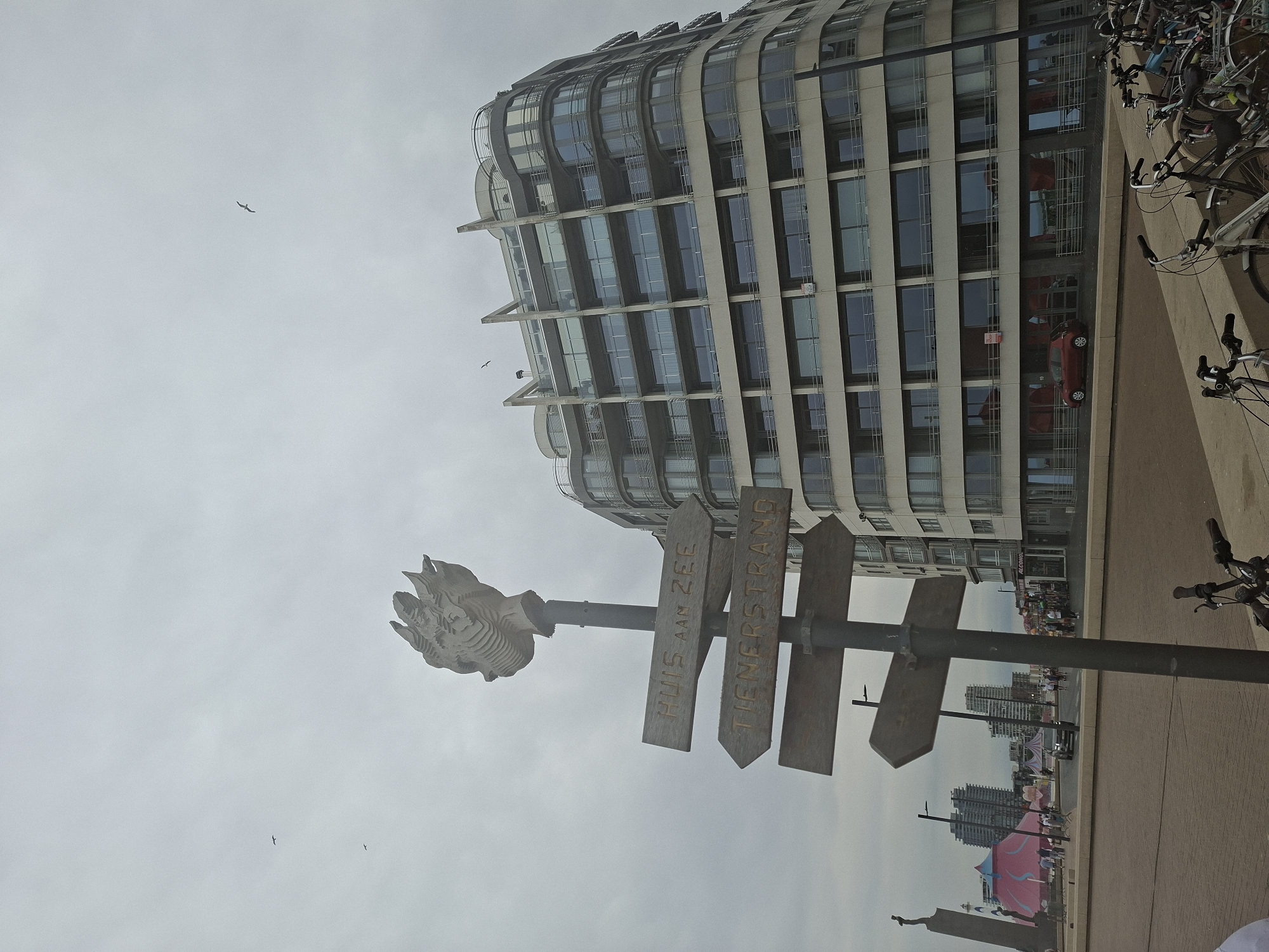 In Ostend, Belgium, on a cloudy day, a directional signpost stands prominently in the foreground. The signpost has a decorative sculpture resembling a stylized head at its apex and features three wooden signs pointing in different directions. The uppermost sign reads "HUIS AAN ZEE", the middle sign reads "TIENERSTRAND", and the bottom sign is blank. Behind the signpost, a modern, multi-story building with a curved facade and numerous windows is visible. In the background, a Ferris wheel and a large pink tent suggest a fairground or event space. Several bicycles are parked in the foreground, and people are walking along a paved promenade. A red car is parked near the building. Birds are seen flying in the overcast sky.