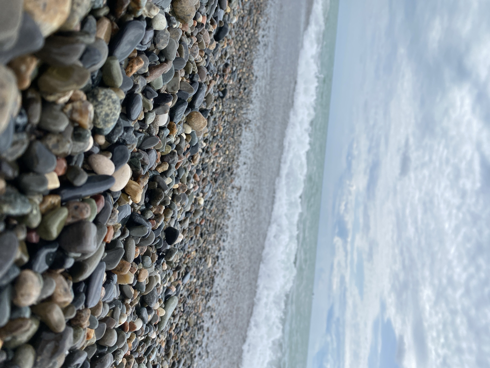 A low-angle, full shot captures a close-up view of a rocky beach in Sokhumi, Abkhazia. The foreground is dominated by a dense collection of smooth, wet pebbles in various shades of gray, brown, black, and white. The pebbles are varied in size and shape, with some glistening from the receding tide. Beyond the pebbles, the turquoise sea stretches to the horizon, with gentle waves breaking white and foamy on the shore. The sky above is overcast with soft, diffused light, casting a serene and slightly moody atmosphere over the scene. The overall impression is one of natural tranquility.