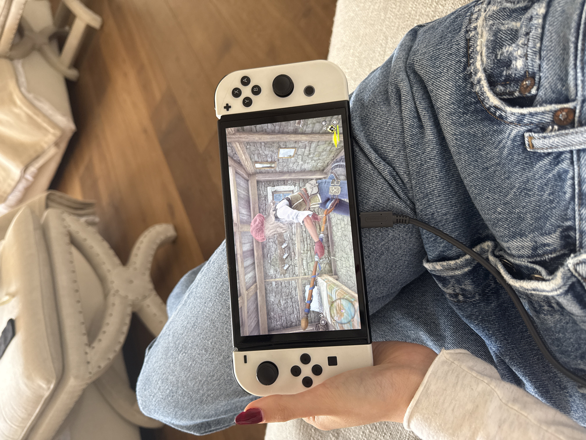 A person wearing jeans is holding a white Nintendo Switch. The game being played appears to be a first-person adventure game set in a medieval-style environment. A charging cable is plugged into the bottom of the device. Part of a chair and a wooden floor are visible in the background. The location is not evident.