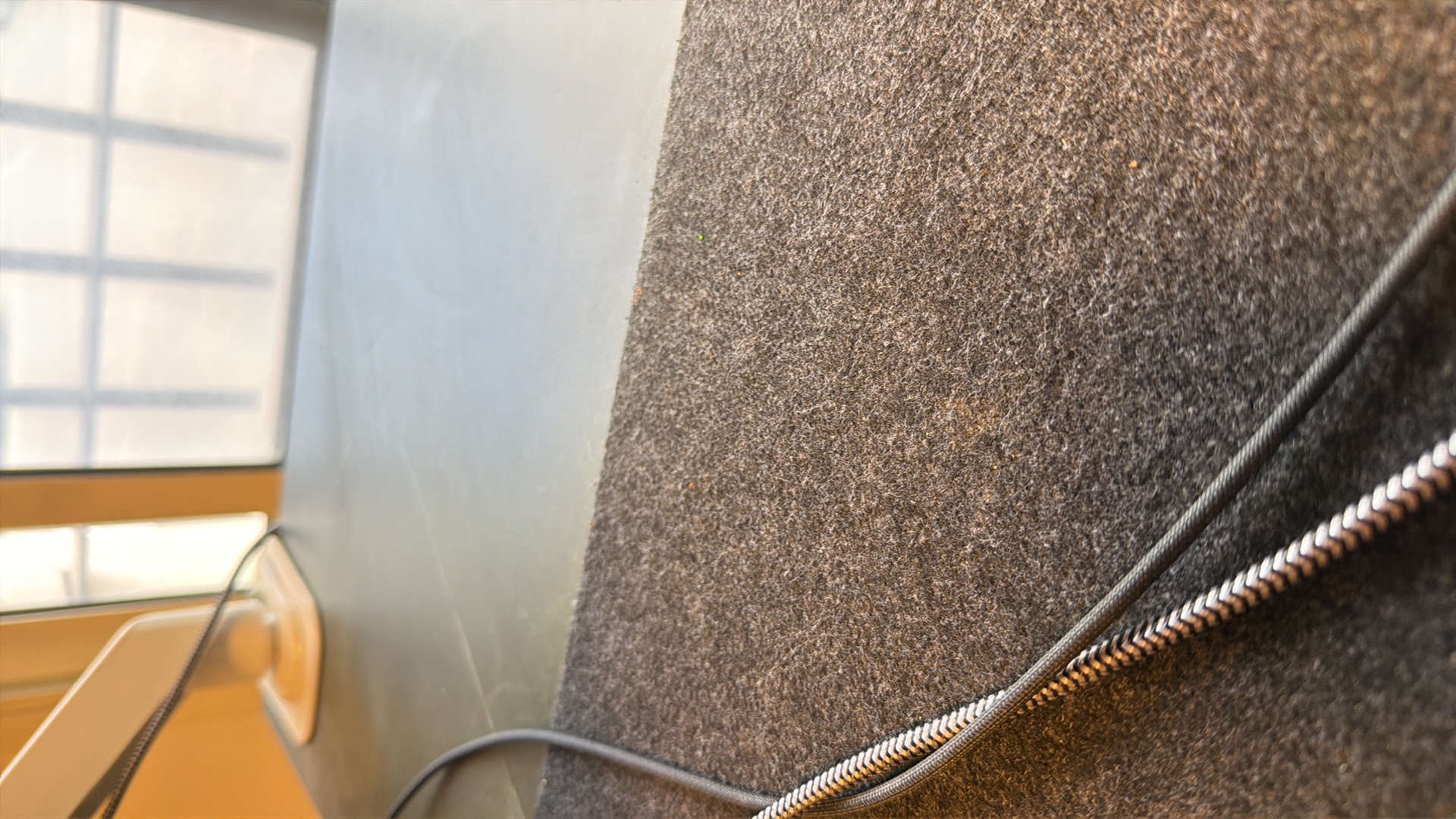 This is a close-up, downward-angled shot of a dark gray carpeted surface with two dark cables running diagonally across it. The cable closest to the viewer has a black and white zigzag pattern, while the cable further away is a solid dark gray. The carpet appears to have some texture and slight variations in color, possibly from dust or wear. In the upper left of the frame, a white monitor arm base is partially visible, suggesting a desk environment. The background shows a blurred window with faint light filtering through, indicating an indoor setting, possibly a home or office. The scene is devoid of any people or specific landmarks that would identify its location within Kafr Yasif, Israel.