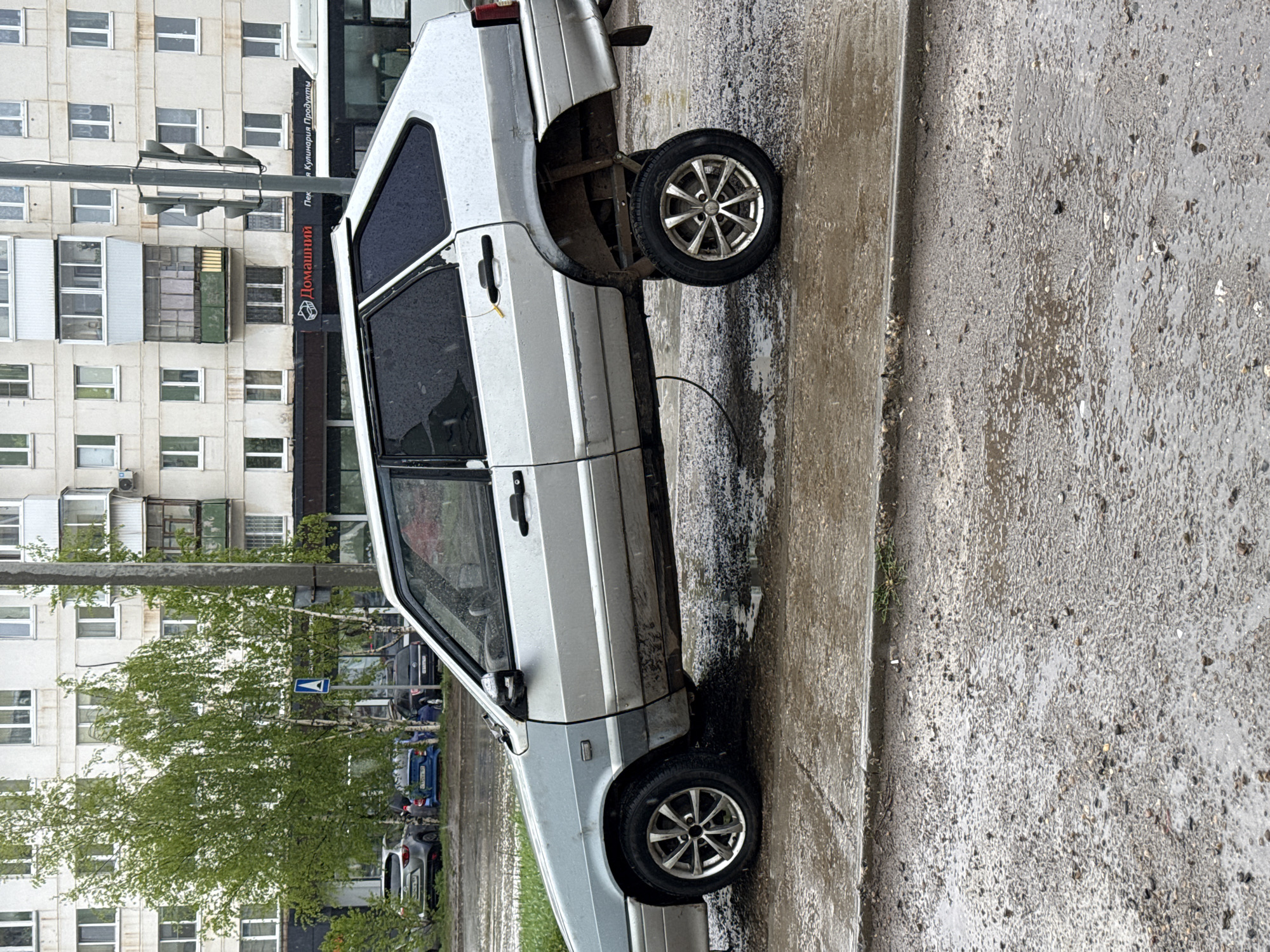 A silver Lada hatchback sits on a flat tire on a wet street in Chaikovsky, Russia. The car is near a curb, and a multistory building is in the background with a sign in Russian reading "Домашний Пекарня Кулинария Продукты". Trees are visible near the building and other cars are parked nearby.