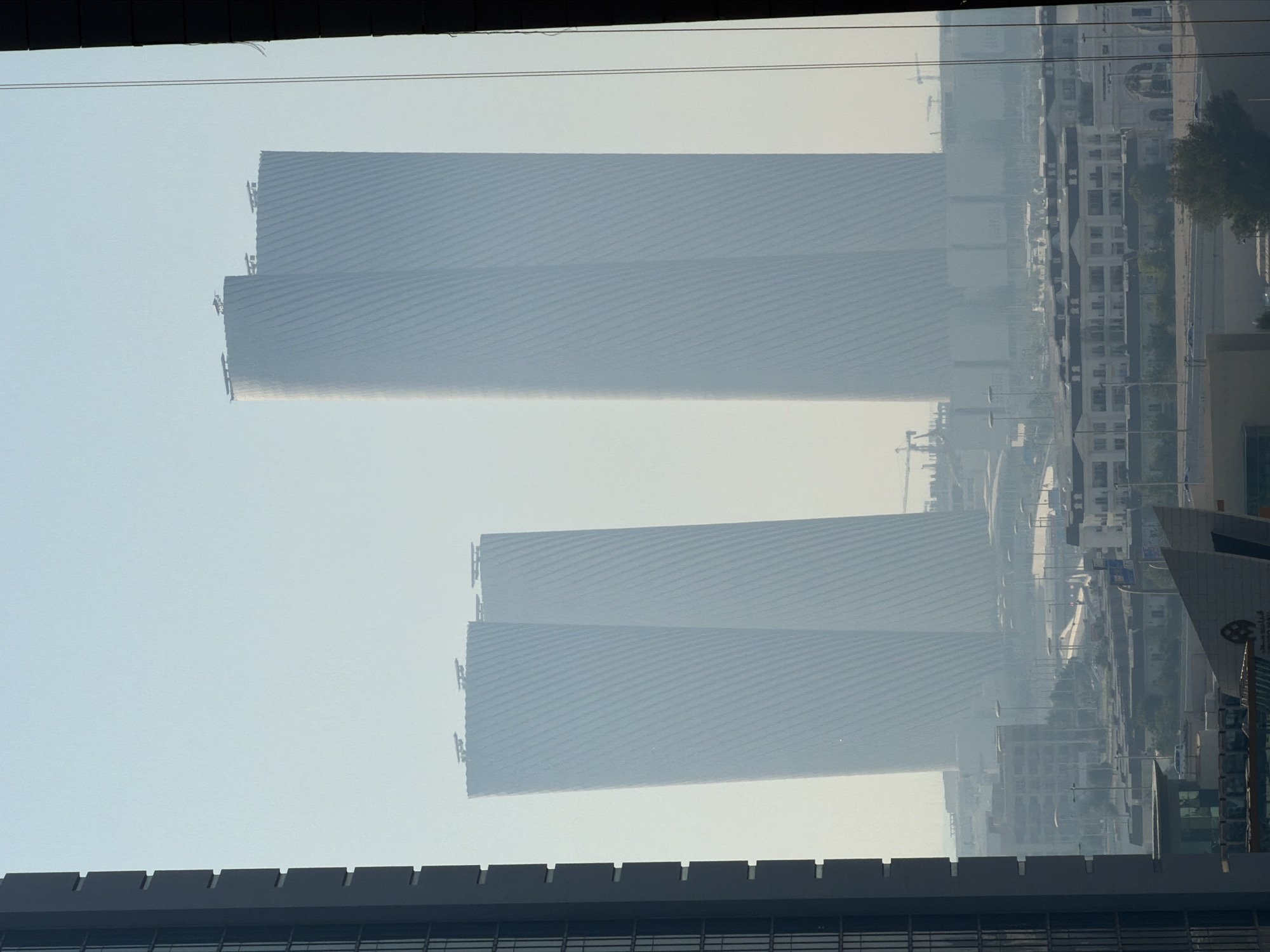 A hazy, high-angle view captures two distinctive, tall, cylindrical buildings in the distance, their facades adorned with diagonal ribbing. These structures appear to be under construction or recently completed, with cranes visible behind them. In the foreground, a dark, modern skyscraper dominates the left side of the frame, its vertical lines and segmented windows suggesting a contemporary architectural style. The middle ground is populated by a dense urban landscape featuring lower-rise buildings with white facades and dark roofs, resembling residential or commercial complexes. Numerous streetlights and roads indicate an active city environment. The overall atmosphere is softened by a light haze or fog, which partially obscures distant details and creates a muted color palette dominated by blues and grays.