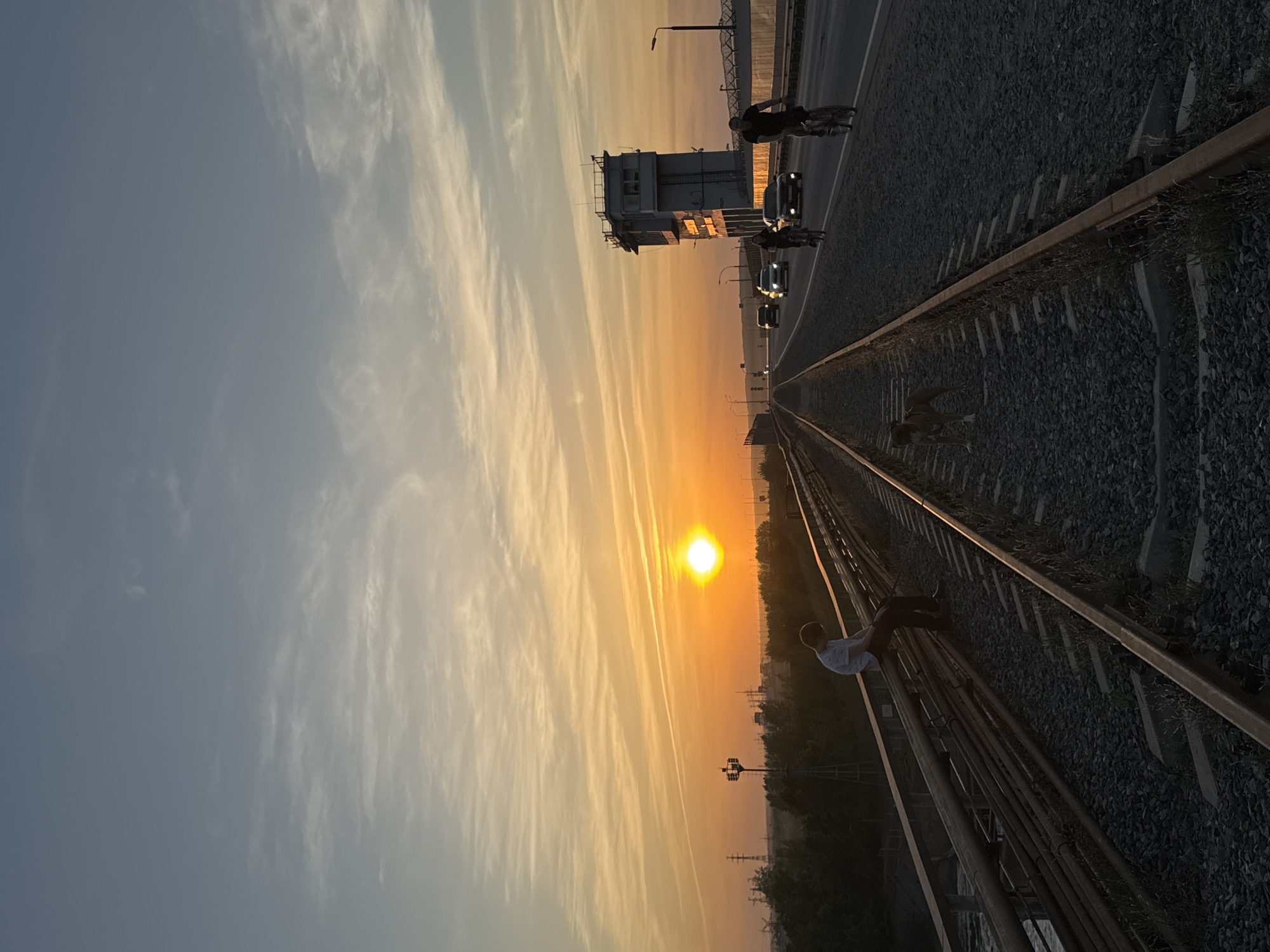 The image depicts a sunset over a railway in Chaikovsky, Russia. The sun is centered in the background, casting an orange glow on the sky and horizon. A railway track extends into the distance, with a person sitting on the side of the rails in the lower right corner. On the left of the frame, there is a road with cars and a cyclist traveling. An electrical transformer box stands beside the road. The image is rotated counterclockwise.