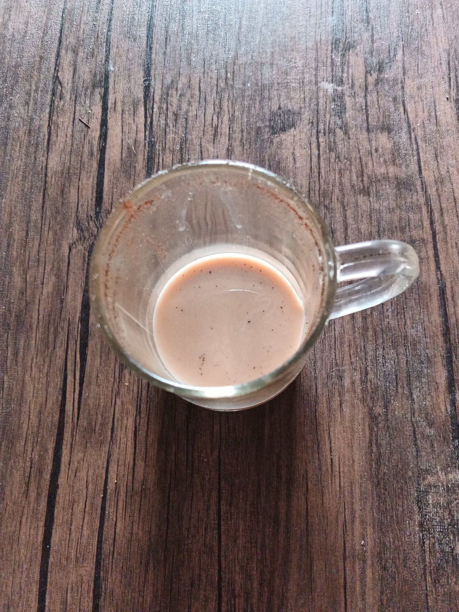 A clear glass mug filled with a light brown liquid, possibly tea or coffee with milk, sits on a dark brown wooden surface. The mug has a clear handle. The wooden surface has a visible grain pattern, with some vertical lines suggesting planks. The liquid inside the mug has small dark specks, likely tea leaves or coffee grounds. The lighting suggests an indoor setting, possibly a home or cafe, with light casting a shadow from the mug onto the surface. There are no people or animals visible in the image. The scene is static and appears to be an everyday object placed on a table. The specific location in Gandasari, Indonesia, cannot be determined from the image alone.