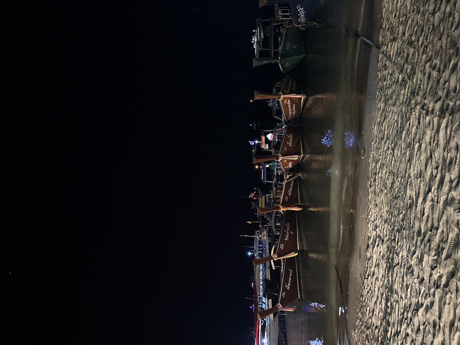 The image depicts a nighttime scene along the shoreline of Ban Ko Li Pe, Thailand. In the foreground, light-colored sand exhibits distinct ripple patterns and faint indentations, indicating a wet surface. The midground is dominated by a fleet of approximately eight boats moored in calm water near the shore. Prominently featured are at least five traditional Thai longtail boats, constructed from dark wood, displaying high pointed bows and tall stern posts. These boats bear Thai script on their hulls, likely names or identifiers, and are equipped with small flags (including the Thai national flag) and low-level artificial lights that reflect on the water's surface. A larger, lighter-colored vessel is partially visible on the far left, and a boat with a green hull is situated towards the right of the longtail fleet. The background is a dark, clear night sky, with minimal distant artificial light sources visible above the horizon. No human figures are present.