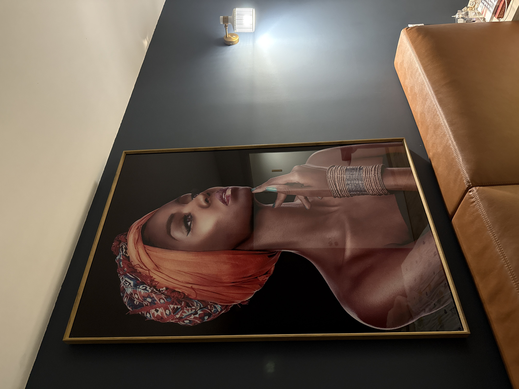 An interior shot depicts a stylish room dominated by a large, vertically oriented framed artwork. The artwork features a portrait of a woman with dark skin, looking upwards and to her right. She wears an elaborate orange and multi-patterned head wrap. Her right hand, adorned with several silver-toned bangles and light blue nail polish, is gently placed near her face. The background of the artwork is solid black, creating a dramatic contrast. The artwork is encased in a slim, gold-colored frame.
The wall behind the artwork is painted in two distinct sections: a dark blue-grey color on the lower part and a cream or off-white color on the upper left, separated by a diagonal line. A modern wall sconce, featuring a gold-colored base and a rectangular, textured glass shade, is mounted on the dark blue-grey wall above the artwork, casting a bright light upwards and downwards.
To the right of the artwork, a segment of a light brown or tan leather sofa is visible, characterized by its smooth texture and clear stitching, suggesting a comfortable and contemporary furniture piece. The scene conveys a sense of modern interior design. No specific activity is taking place, and no external time of day or weather conditions are discernible. There is no legible text visible within the image.