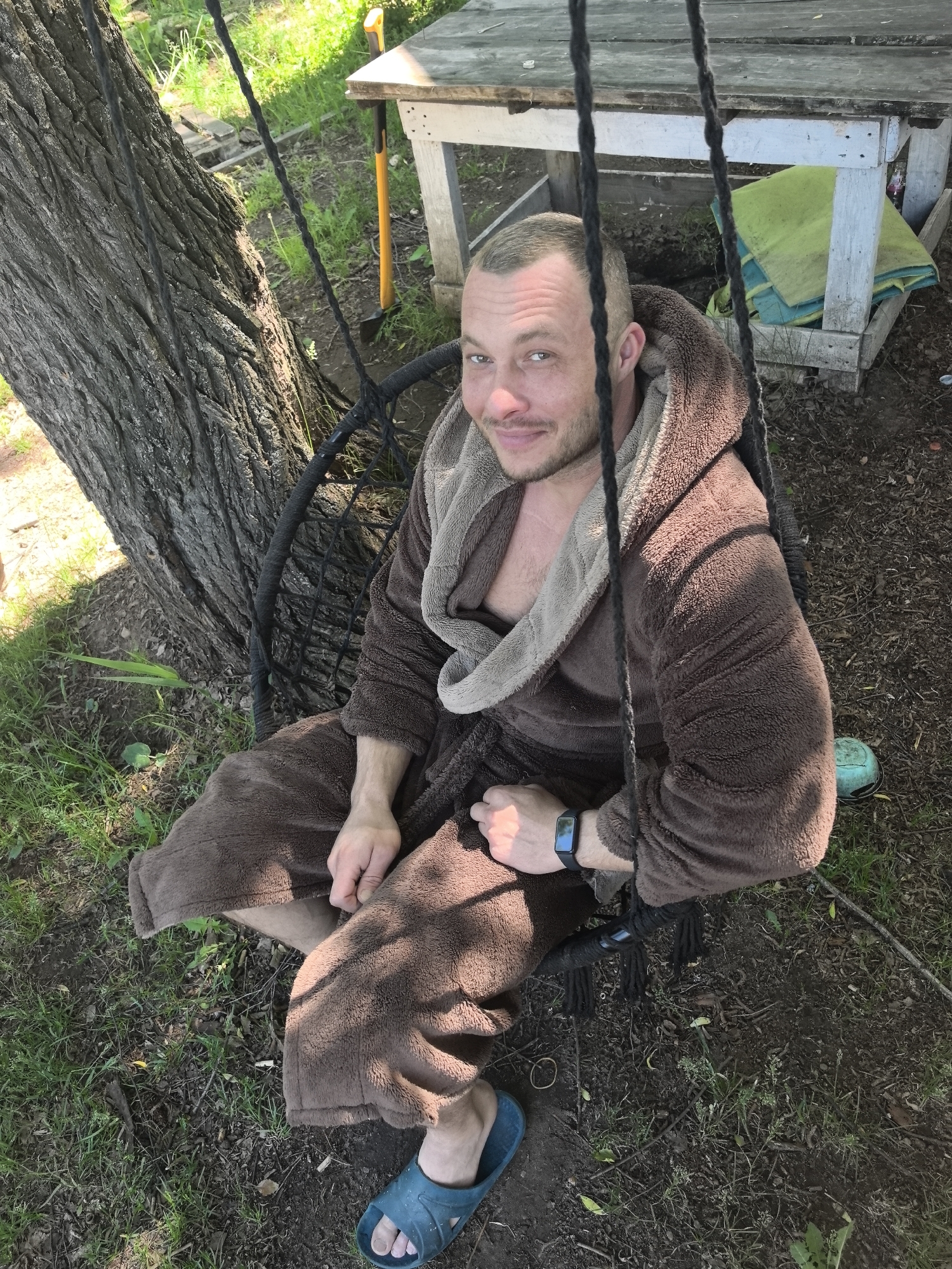 This image depicts a single male subject seated outdoors in a suspended dark-colored swing chair in the city of Noviy, Russia. The man has short, light-colored hair and a light beard, and is wearing a brown, textured hooded bathrobe with a lighter brown or beige lining. A dark-colored fitness tracker or smartwatch is visible on his left wrist. His right foot, wearing a blue rubber slide sandal, is visible in the lower right foreground. He is smiling faintly and looking directly towards the camera.
The swing chair is composed of dark, woven or braided material, suspended by dark ropes. To the left, a large tree with dark, rough bark stands, anchoring the swing. The ground beneath the tree and in the immediate foreground is a mixture of green grass and exposed soil with scattered leaf litter. In the background, to the right of the man, a rustic wooden table, partially painted white, is visible. Leaning against this table is a shovel with a bright yellow handle. Beneath the table, a folded green textile, resembling a towel or blanket, rests on the ground. The environment suggests a casual, outdoor setting, possibly a yard or garden.