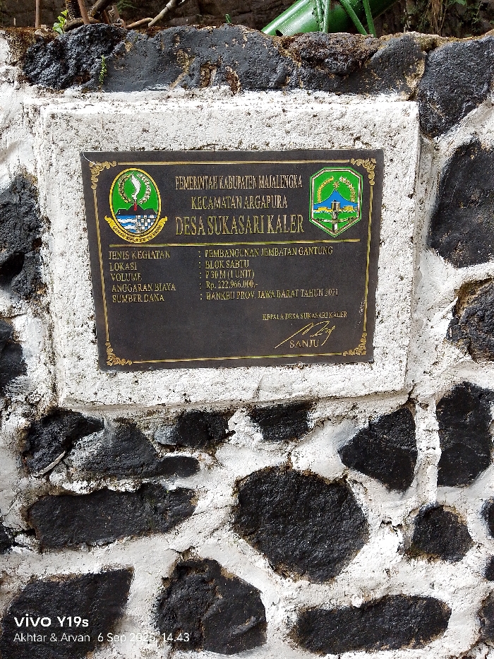 A dark bronze-colored rectangular plaque, framed with a decorative border, is prominently affixed to a wall constructed from irregular, dark-colored stones mortared with white cement. The plaque details a government project in Indonesia.
The top section of the plaque displays "PEMERINTAH KABUPATEN MAJALENGKA" (Government of Majalengka Regency), "KECAMATAN ARGAPURA" (Argapura District), and "DESA SUKASARI KALER" (Sukasari Kaler Village). Flanking these lines are two distinct emblems: the Majalengka Regency emblem on the left and the West Java Provincial Government emblem on the right.
Below, project specifics are listed:
- **JENIS KEGIATAN (Type of Activity):** PEMBANGUNAN JEMBATAN GANTUNG (Suspension Bridge Construction)
- **LOKASI (Location):** BLOK SABTU (Sabtu Block)
- **VOLUME:** P 30 M (1 UNIT) (30 Meters, 1 Unit)
- **ANGGARAN BIAYA (Budget):** Rp. 222.966.000,-
- **SUMBER DANA (Funding Source):** BANKEU PROV. JAWA BARAT TAHUN 2021 (West Java Province Bankeu Year 2021)
The bottom portion of the plaque states "KEPALA DESA SUKASARI KALER" (Head of Sukasari Kaler Village) above a signature, followed by the name "SANJU".
The stone wall beneath the plaque features varying sizes of dark, possibly volcanic, rocks with thick, white mortar joints, some showing minor wear and discoloration. The top edge of the wall where the plaque is situated appears rough, with some dark organic material, possibly dirt or moss, and a sliver of a green object partially visible at the very top of the frame. No people or specific actions are depicted. The image metadata "vivo Y19s Akhtar & Arvan 6 Sep 2023, 14:43" is visible at the bottom left. The location detailed on the plaque is Sukasari Kaler Village, Argapura District, Majalengka Regency, Indonesia.